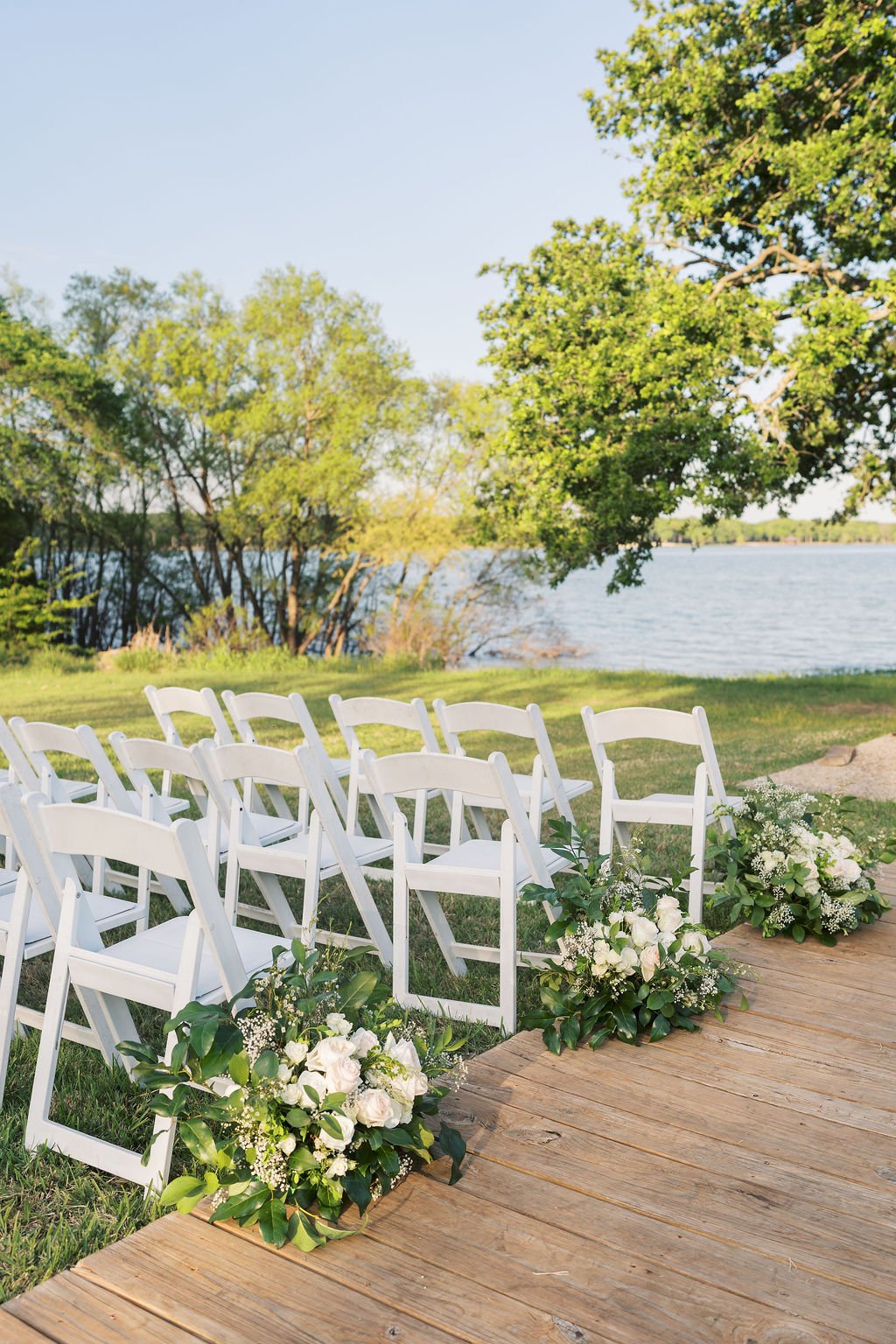 Outdoor wedding ceremony setup by Lake Tawakoni with white chairs and floral arrangements on a wooden aisle.  Hosted at Peninsula Ranch Wedding Venue in Quinlan Texas