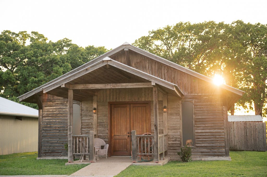A rustic wooden cabin with a front porch, surrounded by green grass and trees, during sunset at Peninsula Ranch in DFW 