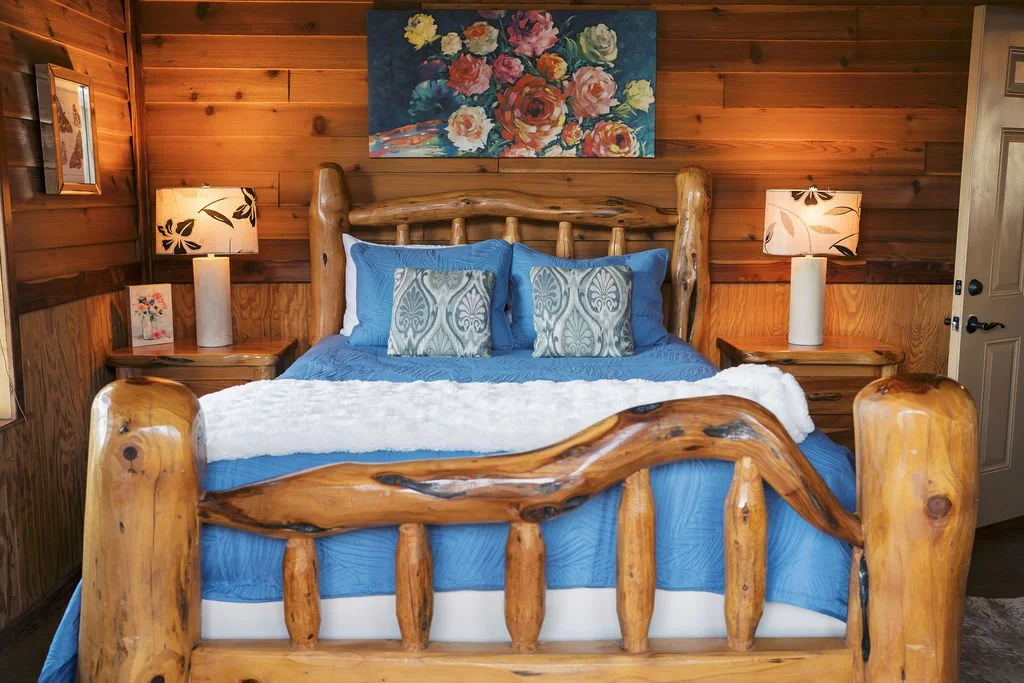 Rustic bedroom with a wooden log bed, blue bedding, decorative pillows, bedside lamps, floral painting, and wood-paneled walls at Peninsula Ranch in DFW 