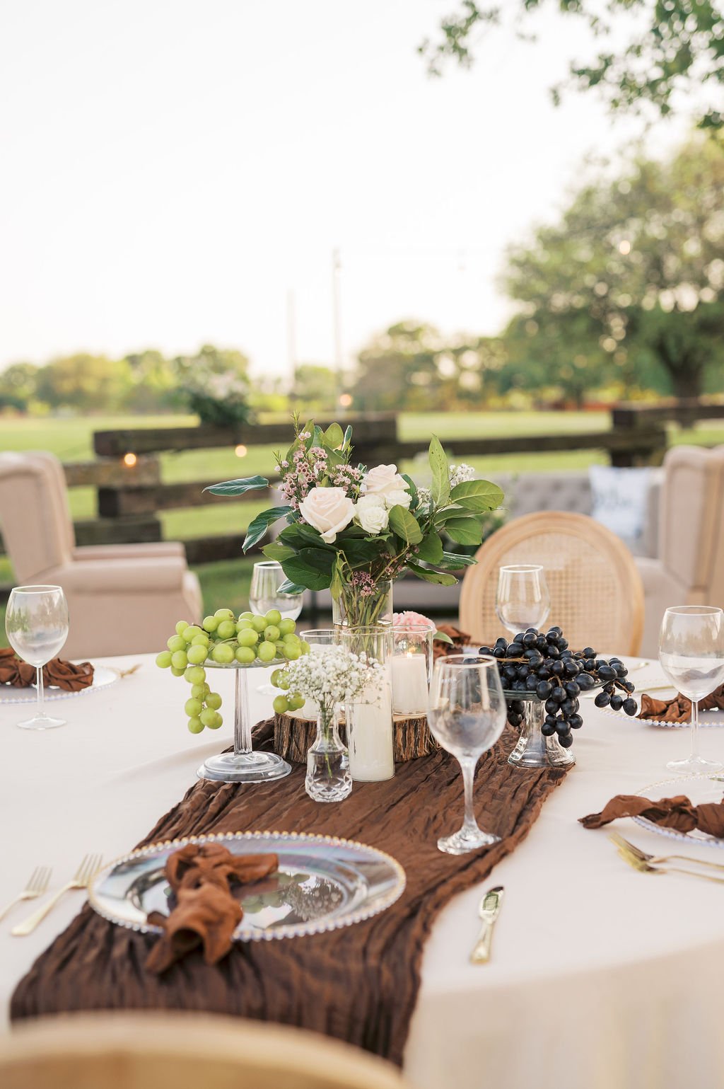 A decorated outdoor dining table at Peninsula Ranch in DFW with a white tablecloth, a brown rustic table runner, and a floral centerpiece in a glass vase. The centerpiece includes white roses, greenery, and small pink flowers. 