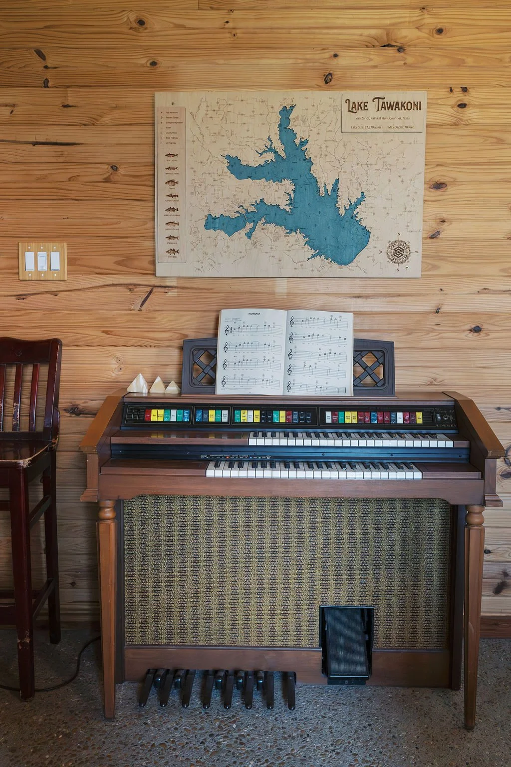 A vintage electric keyboard piano with colorful switches on top, an open sheet music book, and a wooden wall with a map of Lake Tawakoni in Texas hanging above at Peninsula Ranch in DFW 