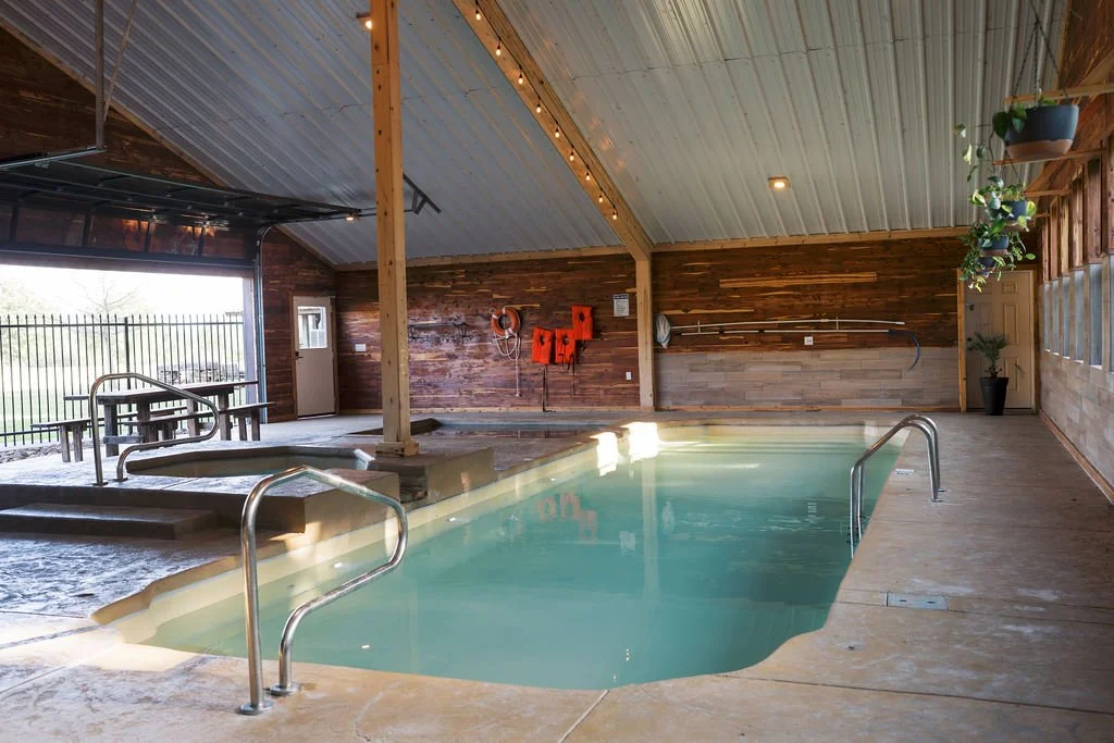 Indoor swimming pool with wooden wall paneling, potted plants, and a sliding door leading outside, with poolside handrails and safety equipment on the wall at Peninsula Ranch in DFW 