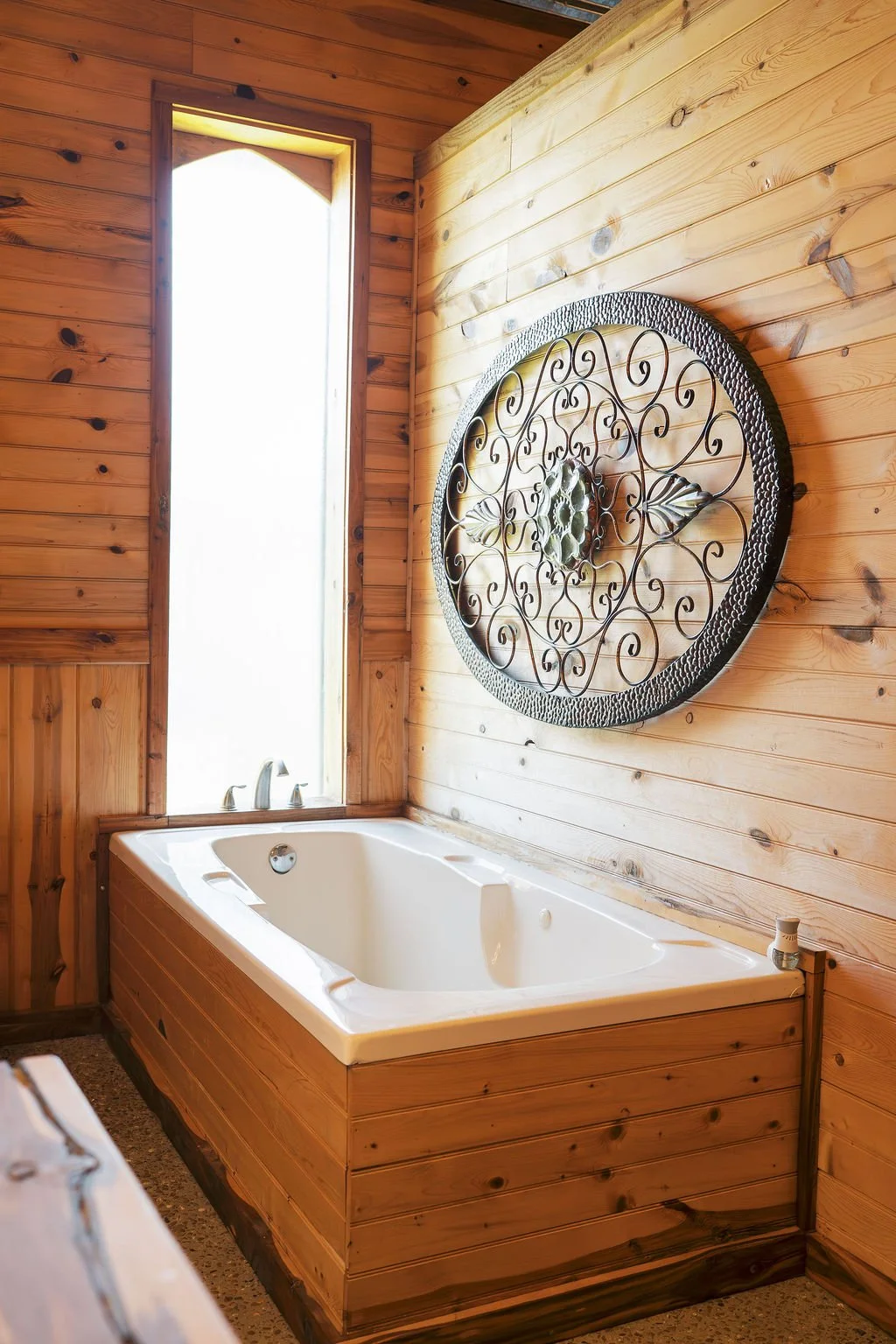 A corner of a bathroom with wooden paneling walls, featuring a white bathtub and a decorative iron wall art piece above it. A tall, narrow window allows natural light to illuminate the space at Peninsula Ranch in DFW 