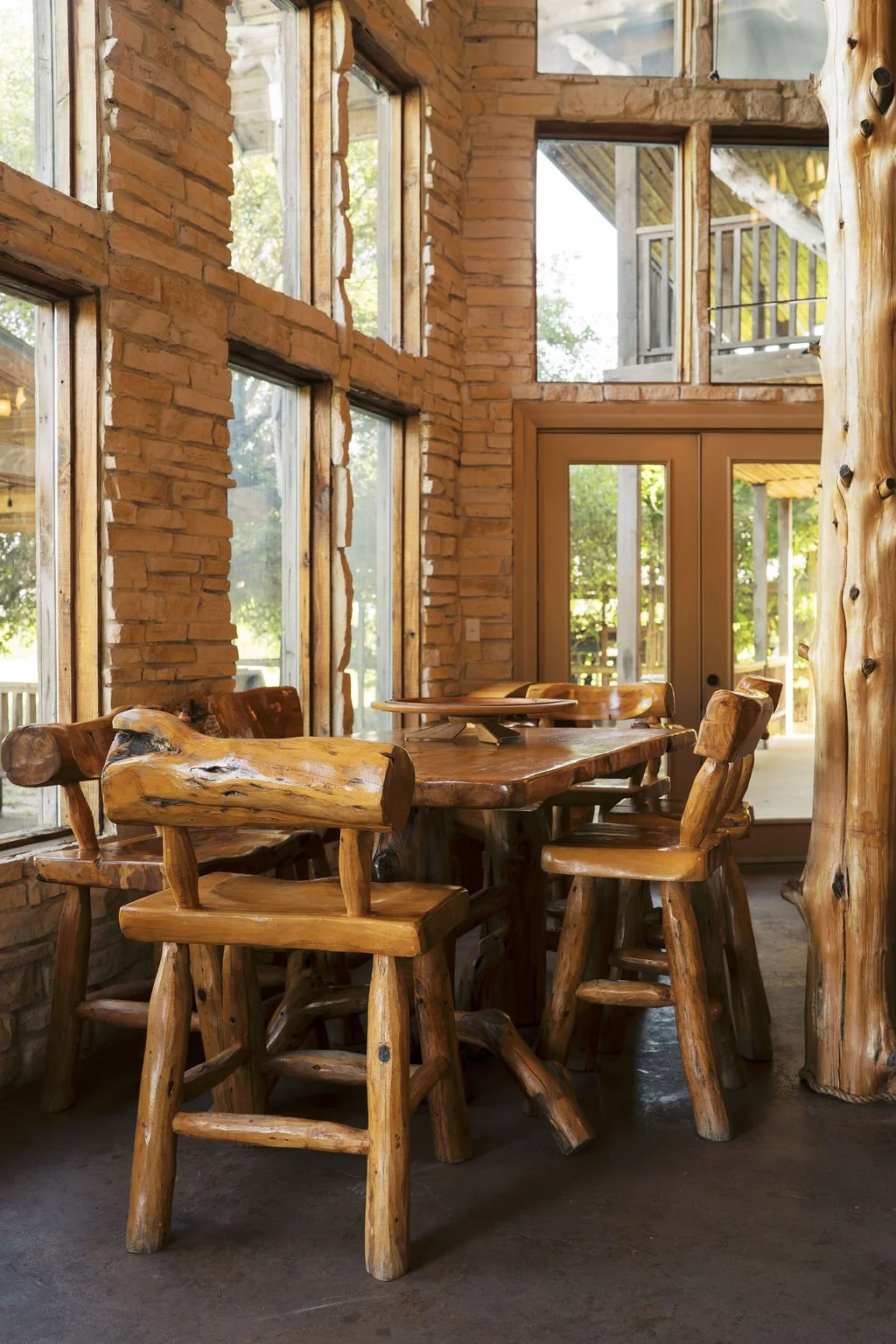 Interior of a rustic dining area of our lodge with a wooden table and matching chairs made from natural logs, surrounded by large windows and brick walls, with a door leading outside at Peninsula Ranch in DFW 
