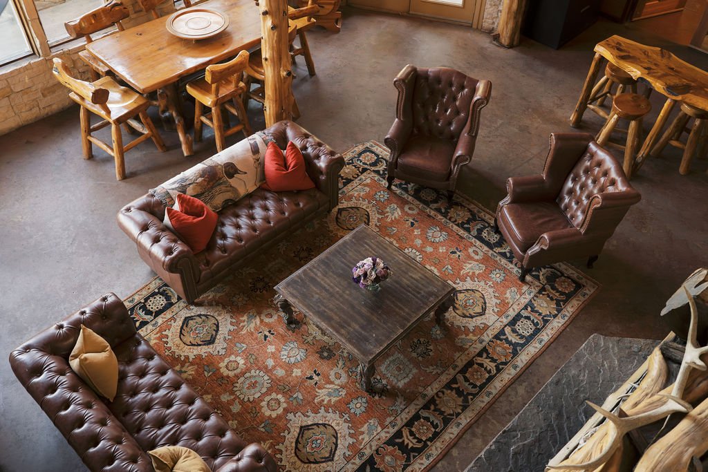Living room with two brown leather tufted sofas, two leather armchairs, a wooden coffee table with a small flower arrangement, an ornate area rug, and a dining area with a wooden table and chairs in the background at Peninsula Ranch in DFW 