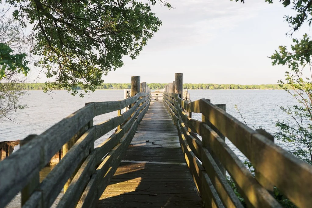 A wooden pier extends over Lake Tawakoni, framed by leafy trees, with a distant shoreline under a cloudy sky at Peninsula Ranch in DFW 