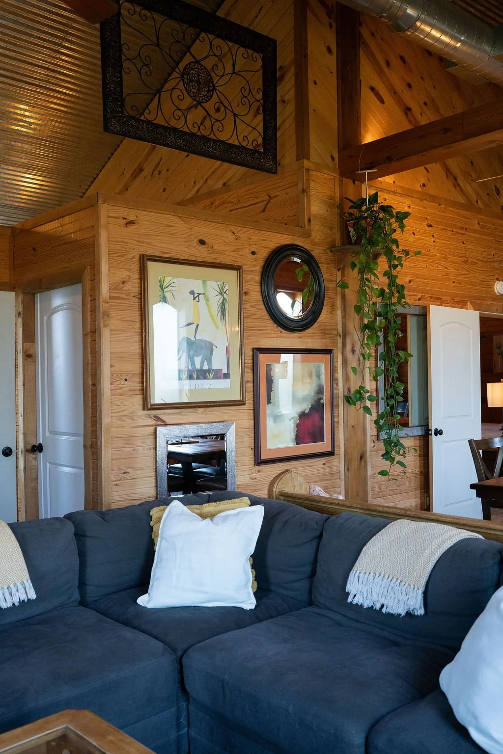 Living room with wood-paneled walls, a gray sectional sofa with white and yellow pillows, wall art including a framed illustration of an animal, a small round mirror, and a hanging plant at Peninsula Ranch in DFW 