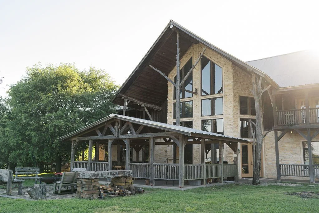 A multi-story hand-built lodge house with large front windows, a covered porch, and a balcony, surrounded by trees and outdoor seating at Peninsula Ranch in DFW.