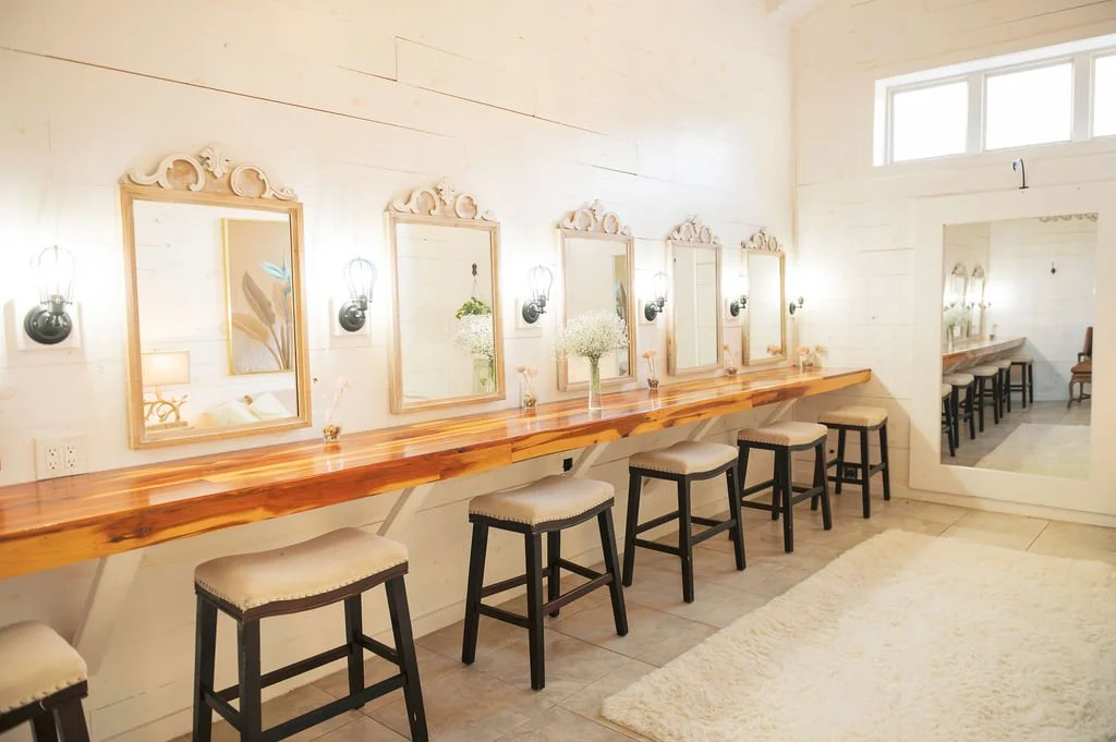 A well-lit dressing room with six mirrors, each with decorative wooden frames, above a long wooden counter with six bar stools, and wall-mounted lights beside each mirror for Brides at Peninsula Ranch in DFW.