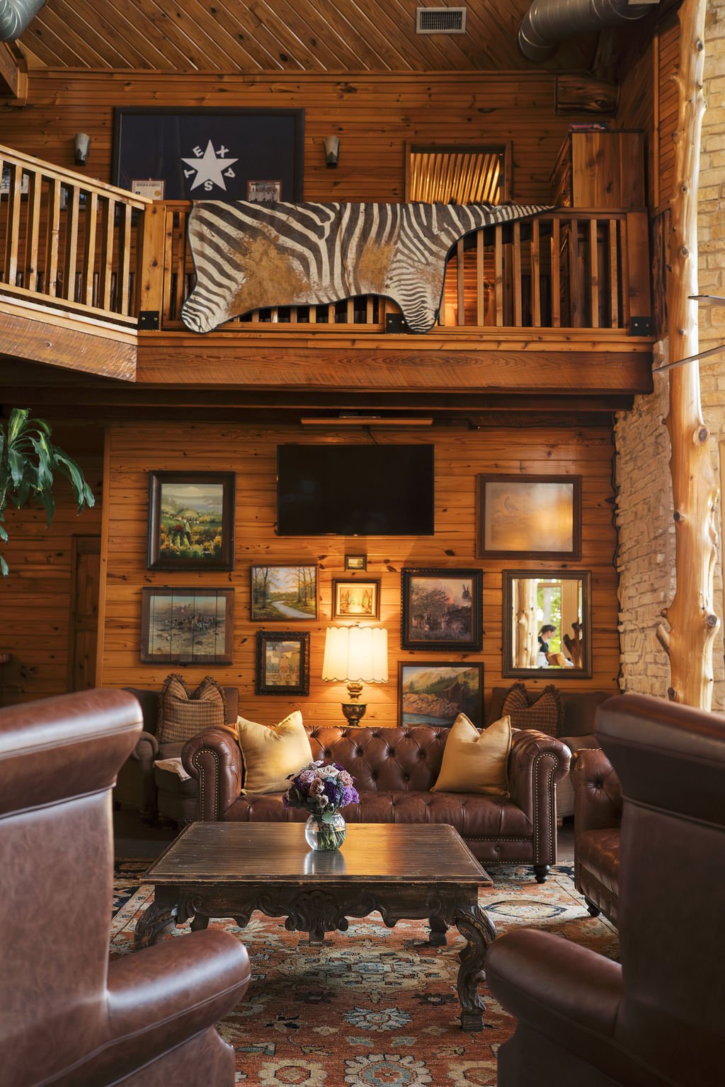 Living room with wooden walls, leather furniture, a wood coffee table with a flower arrangement, wall art, and a TV. A zebra hide decorates the balcony railing at Peninsula Ranch in DFW 