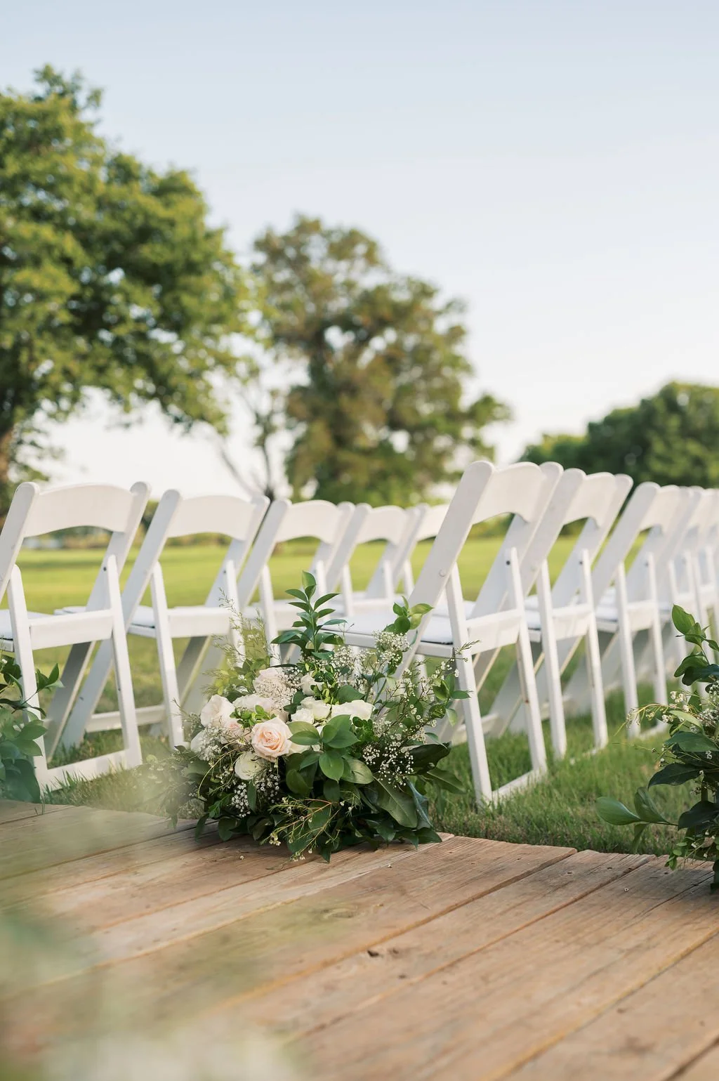 Outdoor wedding ceremony at Peninsula Ranch with white folding chairs arranged in rows, a bouquet of IZALENA Floral House flowers on a wooden platform, green trees in the background, and a clear sky.