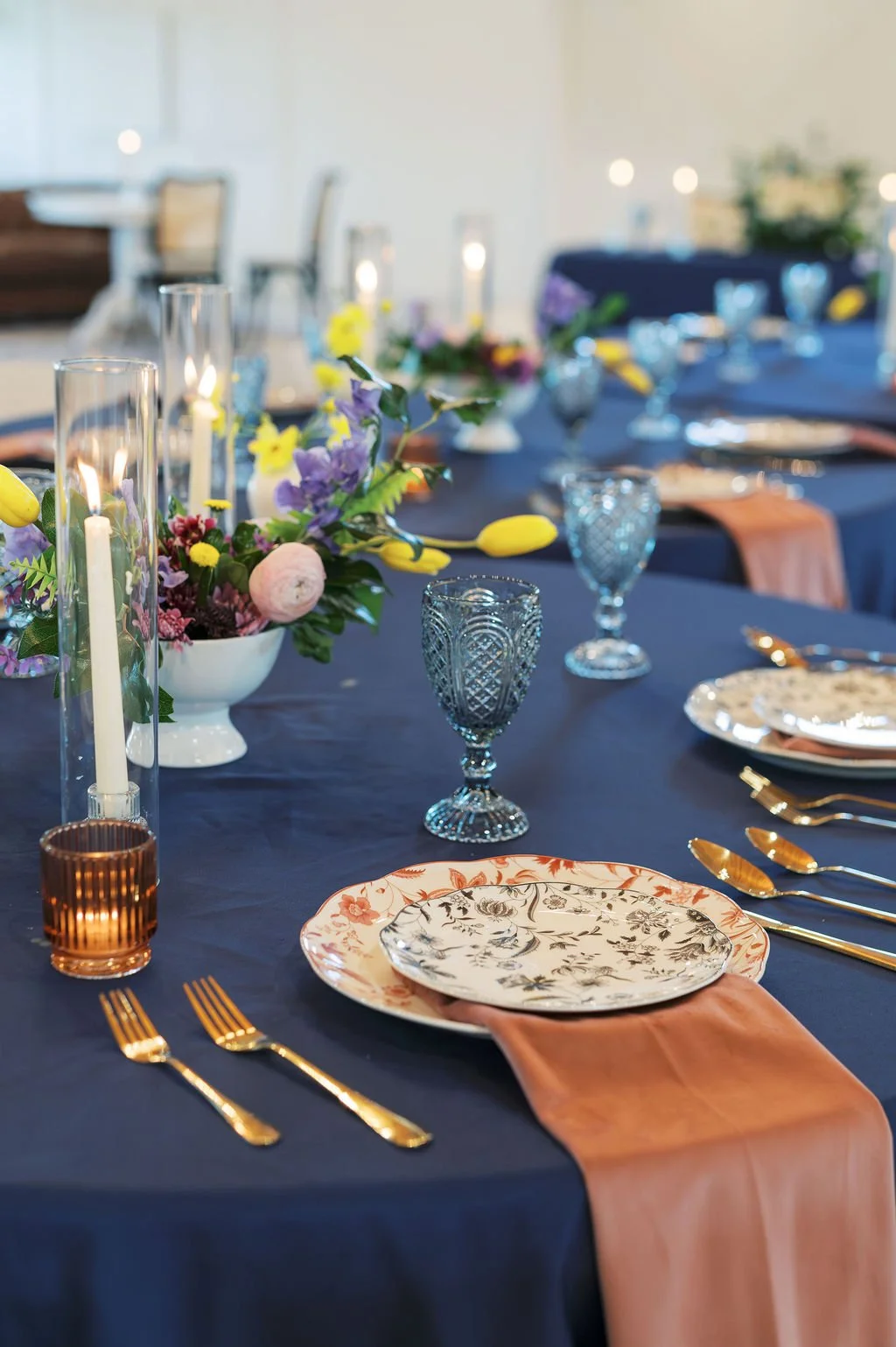Elegant table setting with floral centerpiece, blue tablecloth, gold cutlery, patterned plates, blue glassware, and candles for a wedding reception at Peninsula Ranch in DFW.