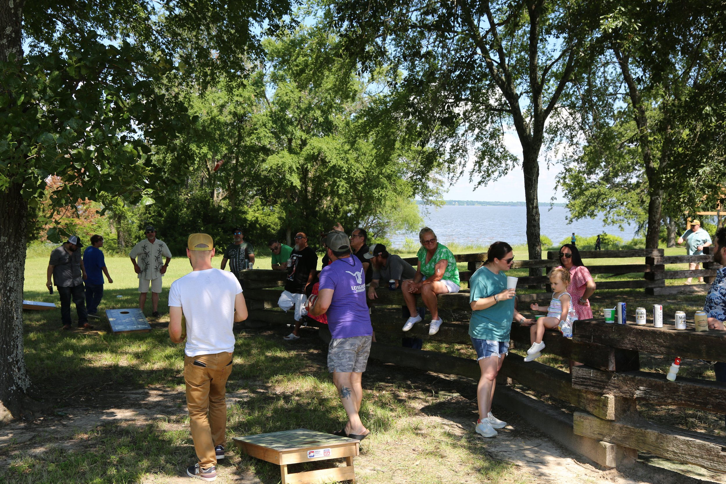 A group of people gathered outdoors near a lake, sitting on wooden benches under trees on a sunny day at Peninsula Ranch in DFW.