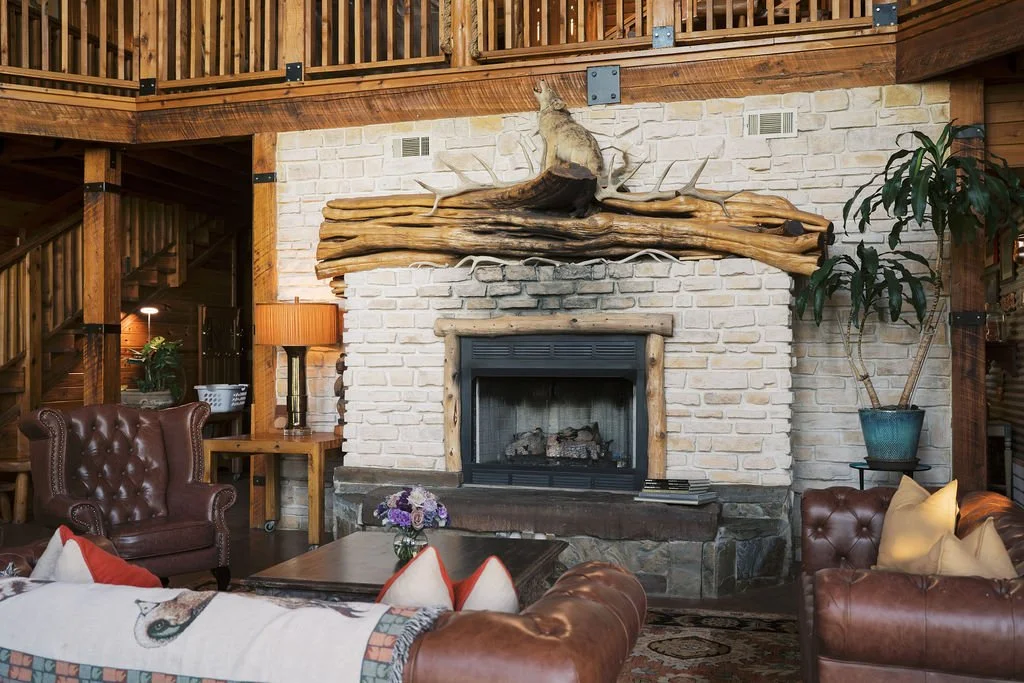 Cozy living room with a white brick fireplace, wooden mantel, and antler decor. Leather armchairs and a sofa with colorful pillows surround a wooden coffee table with a flower arrangement at Peninsula Ranch in DFW 
