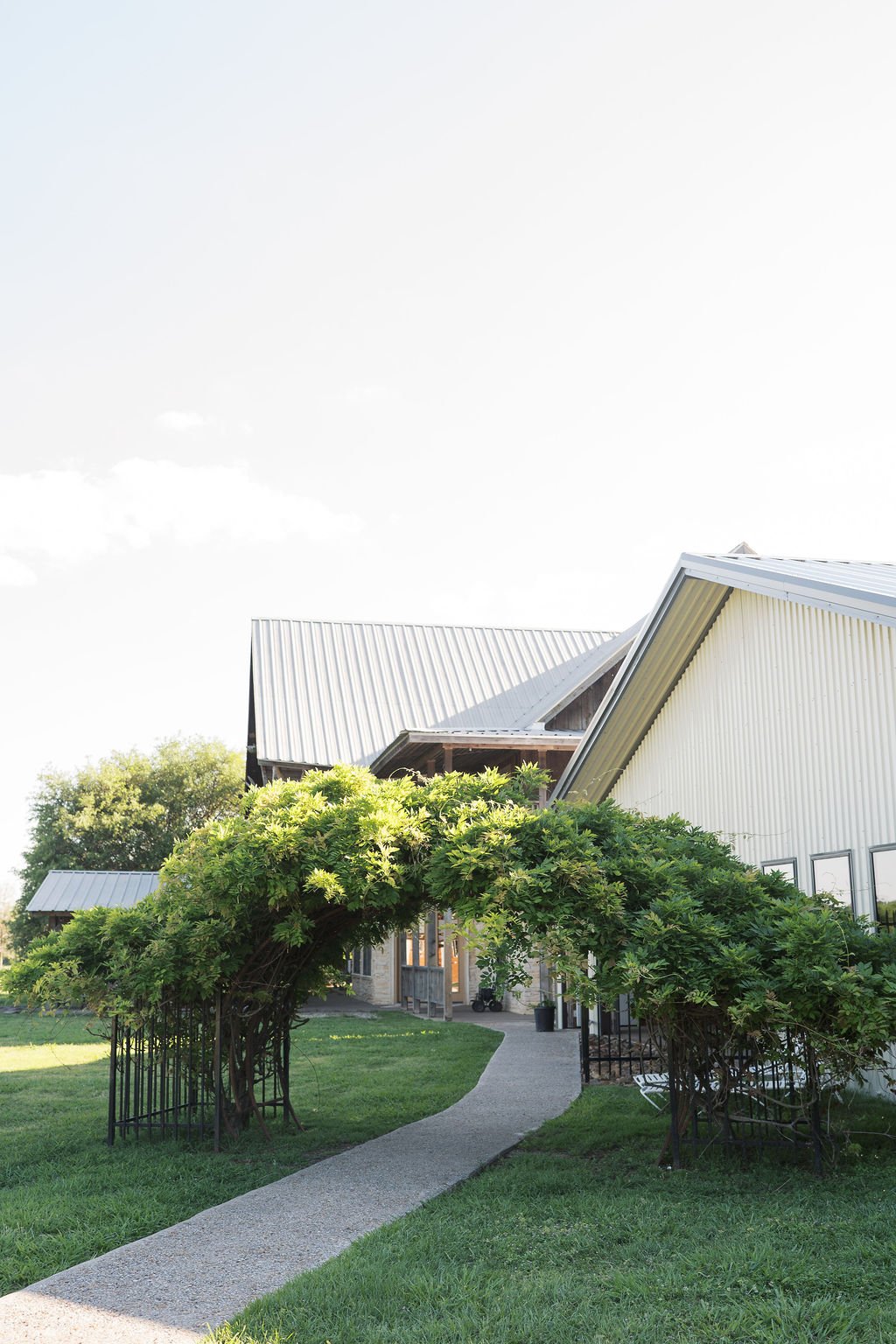A curved wooden archway covered with green foliage leading to a modern house with metal roofs and several windows, surrounded by a grassy yard at Peninsula Ranch in DFW 