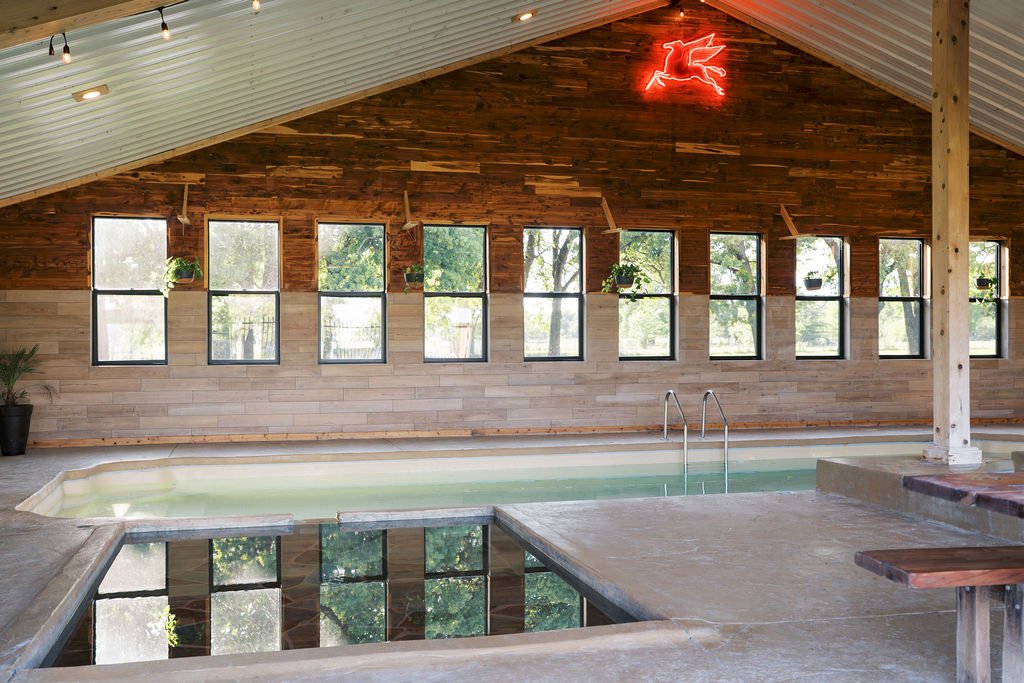 Indoor swimming pool with a curved edge, surrounded by a concrete deck and supported by wooden beams and paneling, with multiple windows on the back wall letting in natural light at Peninsula Ranch in DFW 