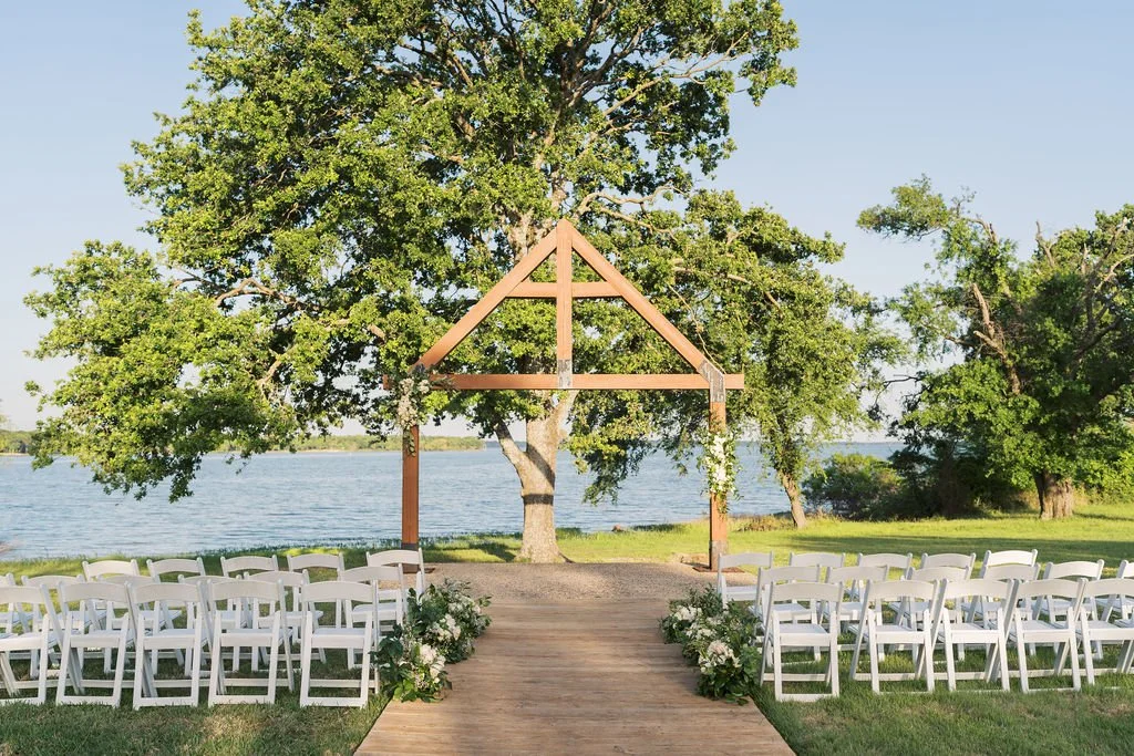 Outdoor wedding ceremony at Peninsula Ranch on Lake Tawakoni with white chairs, wooden aisle, and a wooden arch under a leafy tree.
