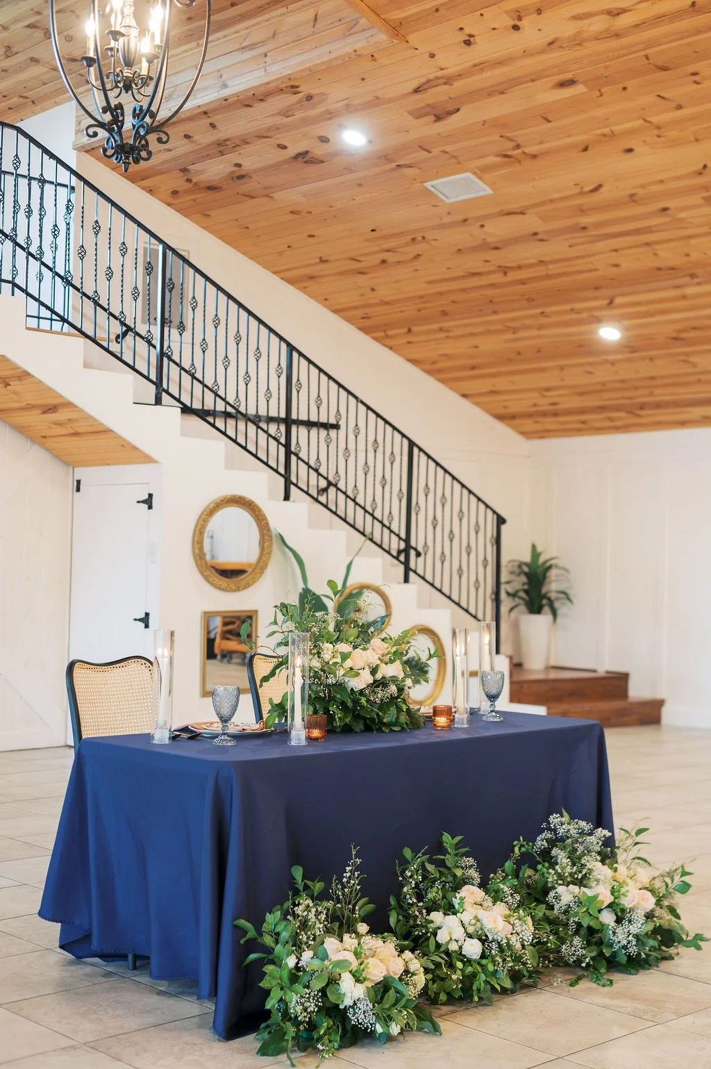 A dining table decorated for an event, covered with a navy tablecloth, featuring IZALENA Floral House floral centerpieces, glassware, and candles, with a wooden ceiling and staircase in the background of the reception hall at Peninsula Ranch in DFW.