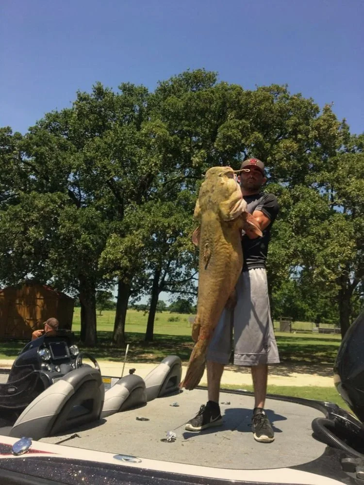 A man standing on a boat holds a very large fish, possibly a catfish, caught in Lake Tawakoni Texas