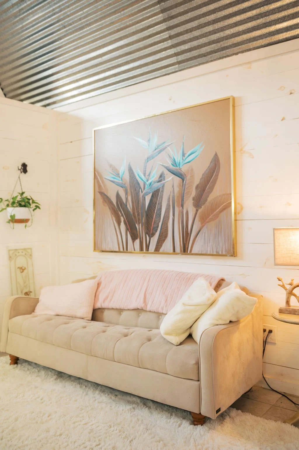 Bridal Suite room with beige tufted sofa, pink and white pillows, a large framed botanical artwork, a hanging plant, a side table with a branch-shaped lamp, and cream-colored walls with a metal ceiling at Peninsula Ranch in DFW.
