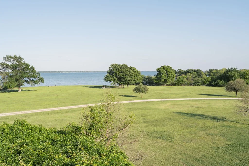 Green open park with scattered trees, dirt path winding through the grass, and Lake Tawakoni in the background beneath a clear blue sky at Peninsula Ranch in DFW 