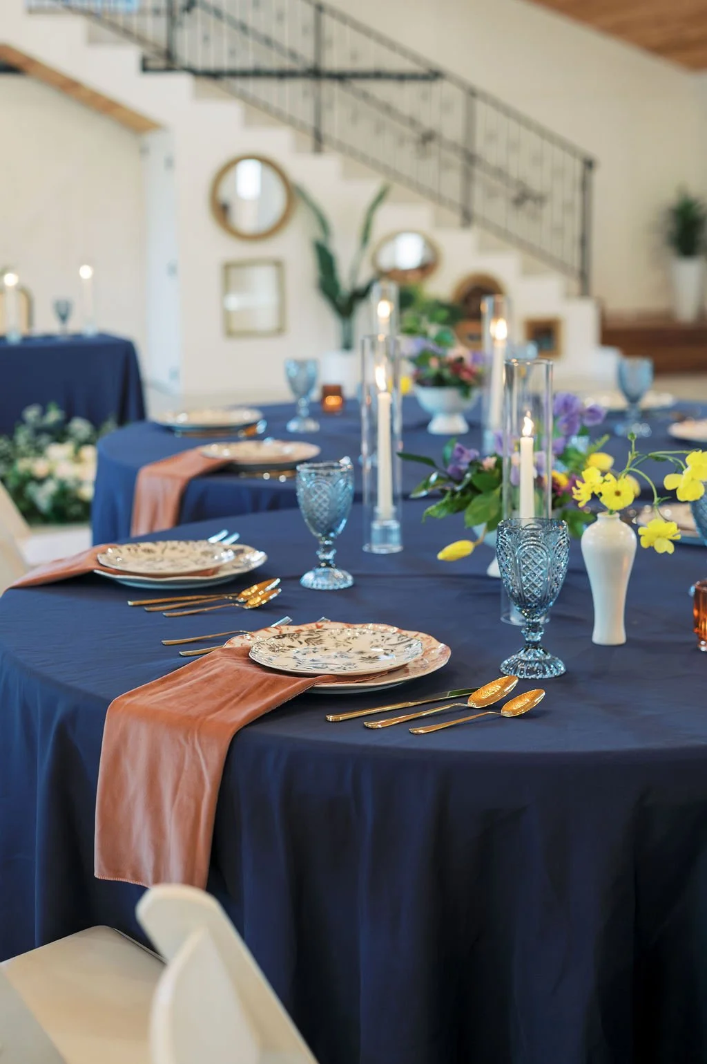 Elegant dining table setup with navy tablecloth, floral plates, gold cutlery, pink napkins, and floral centerpieces with yellow and purple flowers, set in a bright room with staircase and mirrors at Peninsula Ranch Wedding Venue.