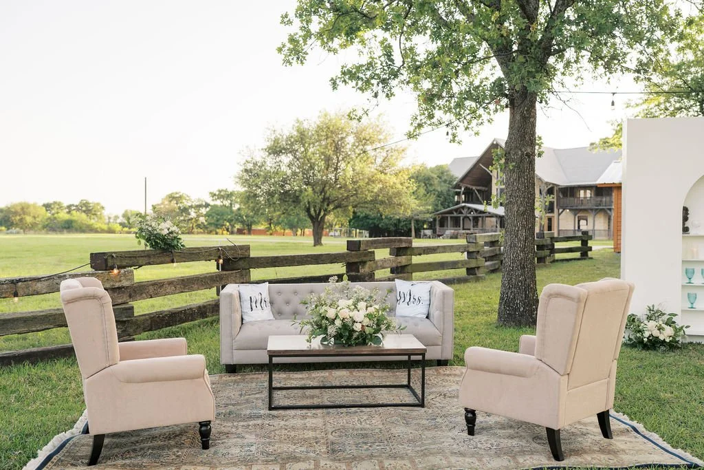 Outdoor reception at Peninsula Ranch Wedding Venue with cream armchairs, a white sofa, and a coffee table with flowers on a grassy lawn, surrounded by trees and wooden fences.