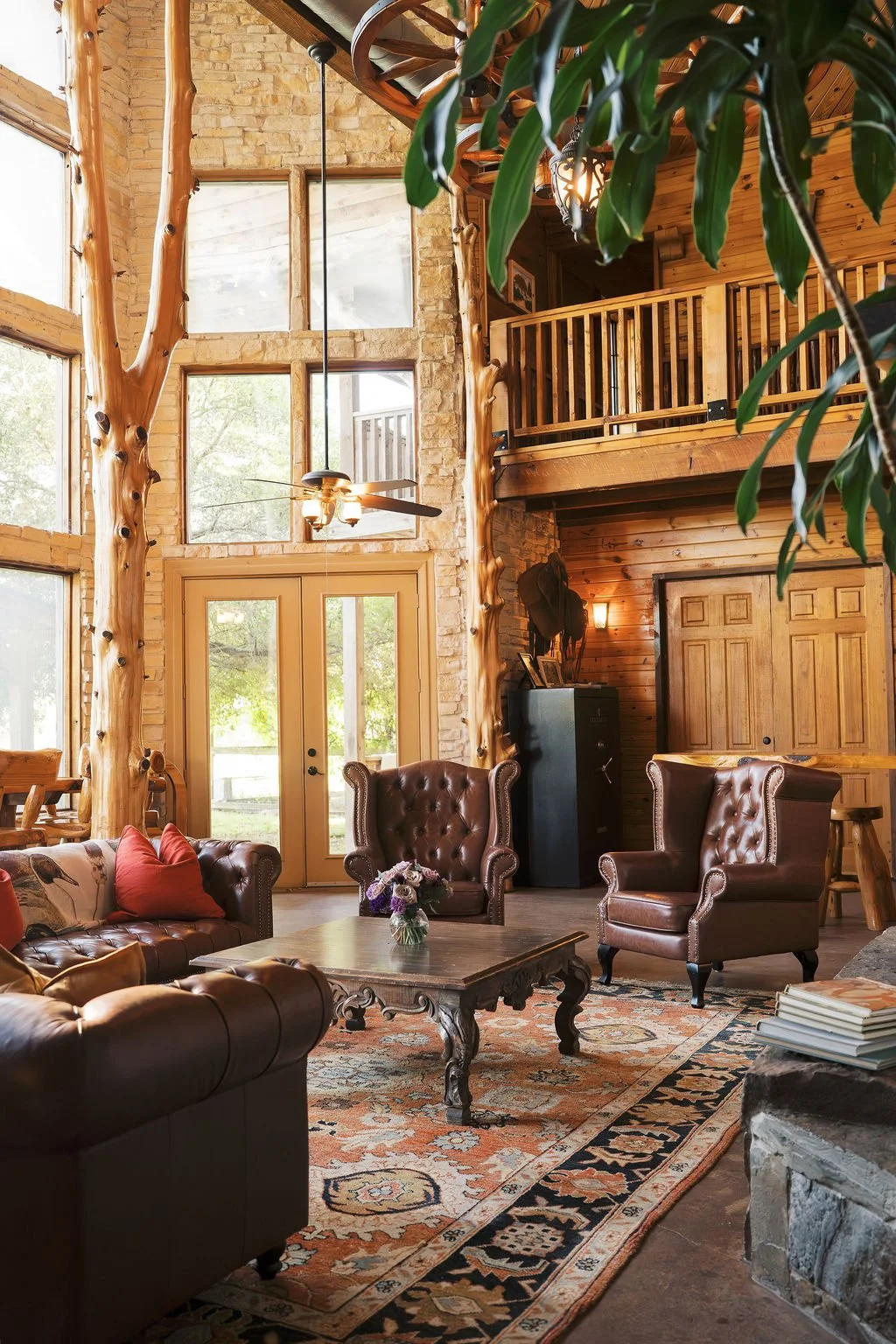 A rustic living room in our lodge with high ceilings, large windows, stone and wood walls, and wooden beams. Features leather armchairs, a wooden coffee table with a flower arrangement, a patterned rug, and a fireplace at Peninsula Ranch in DFW 