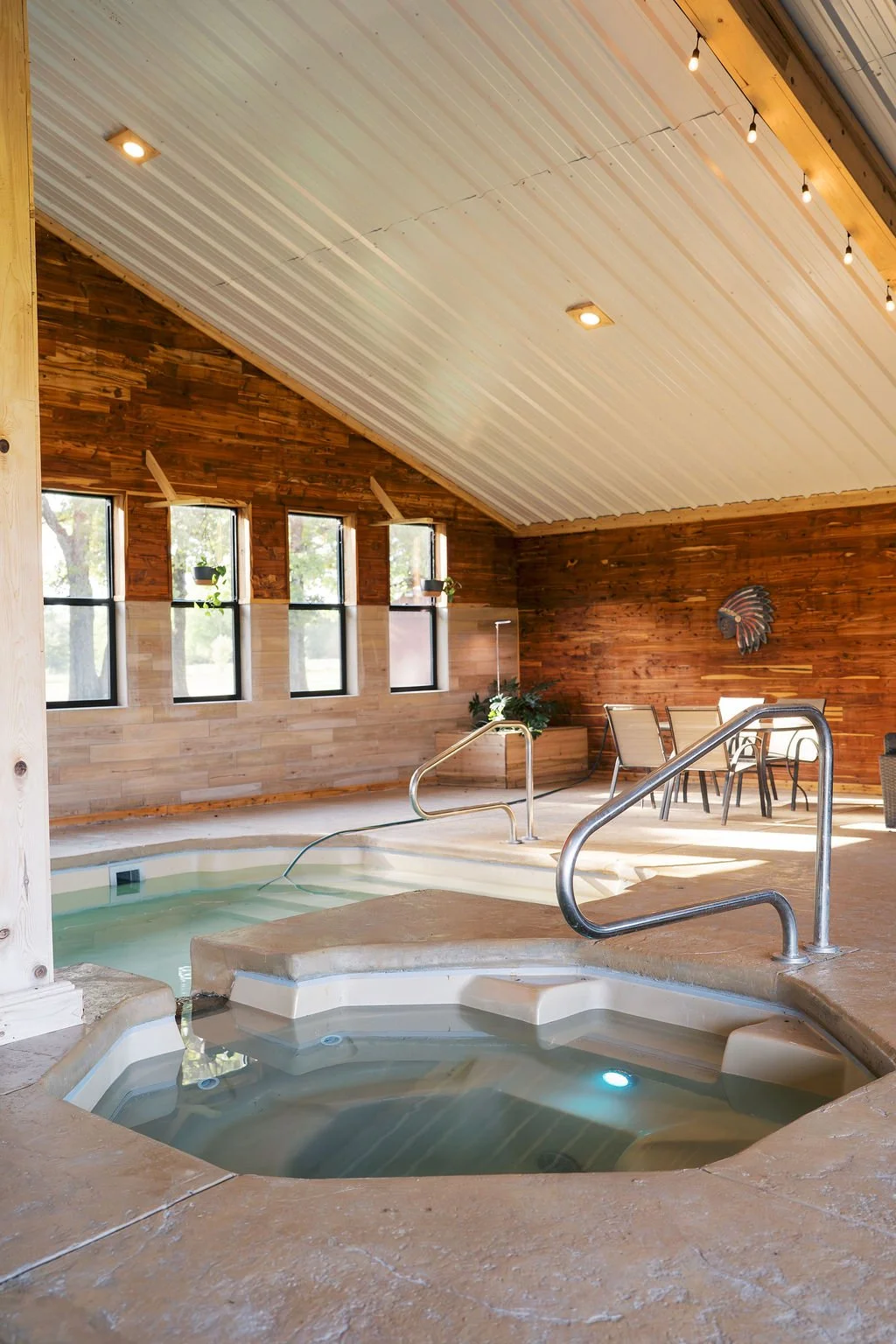 Indoor hot tub at Peninsula Ranch in DFW with multiple seating areas, metal handrails, and a wood-paneled wall with a decorative feather art piece, under a high, sloped ceiling with string lights.