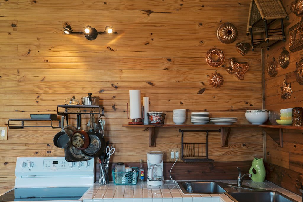 Kitchen with wooden wall, stove, hanging pots and pans, open shelves with dishes and utensils, countertop with coffee maker, sink, and kettle at Peninsula Ranch in DFW 