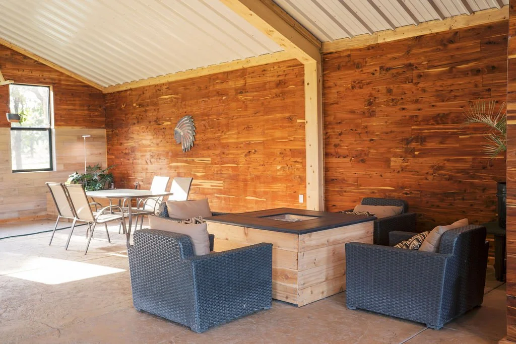 Interior of a living room in our indoor pool with wooden walls, a dining table with chairs, and cushioned armchairs around a firepit table at Peninsula Ranch in DFW 