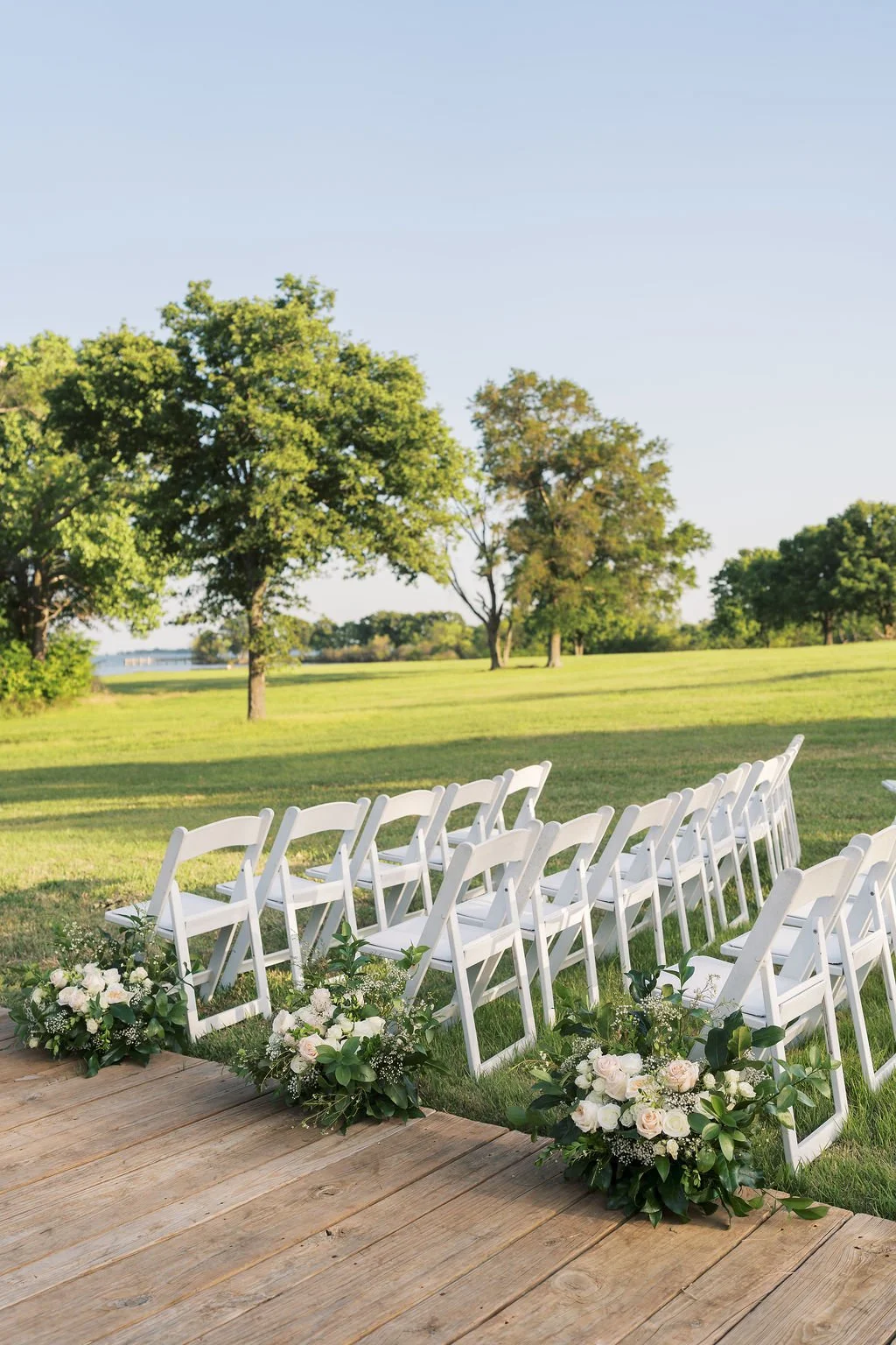 Outdoor ceremony at Peninsula Ranch Wedding Venue with white chairs arranged in rows on a grassy field, decorated with floral arrangements by IZALENA Floral House at the front, against a backdrop of green trees and a clear blue sky.
