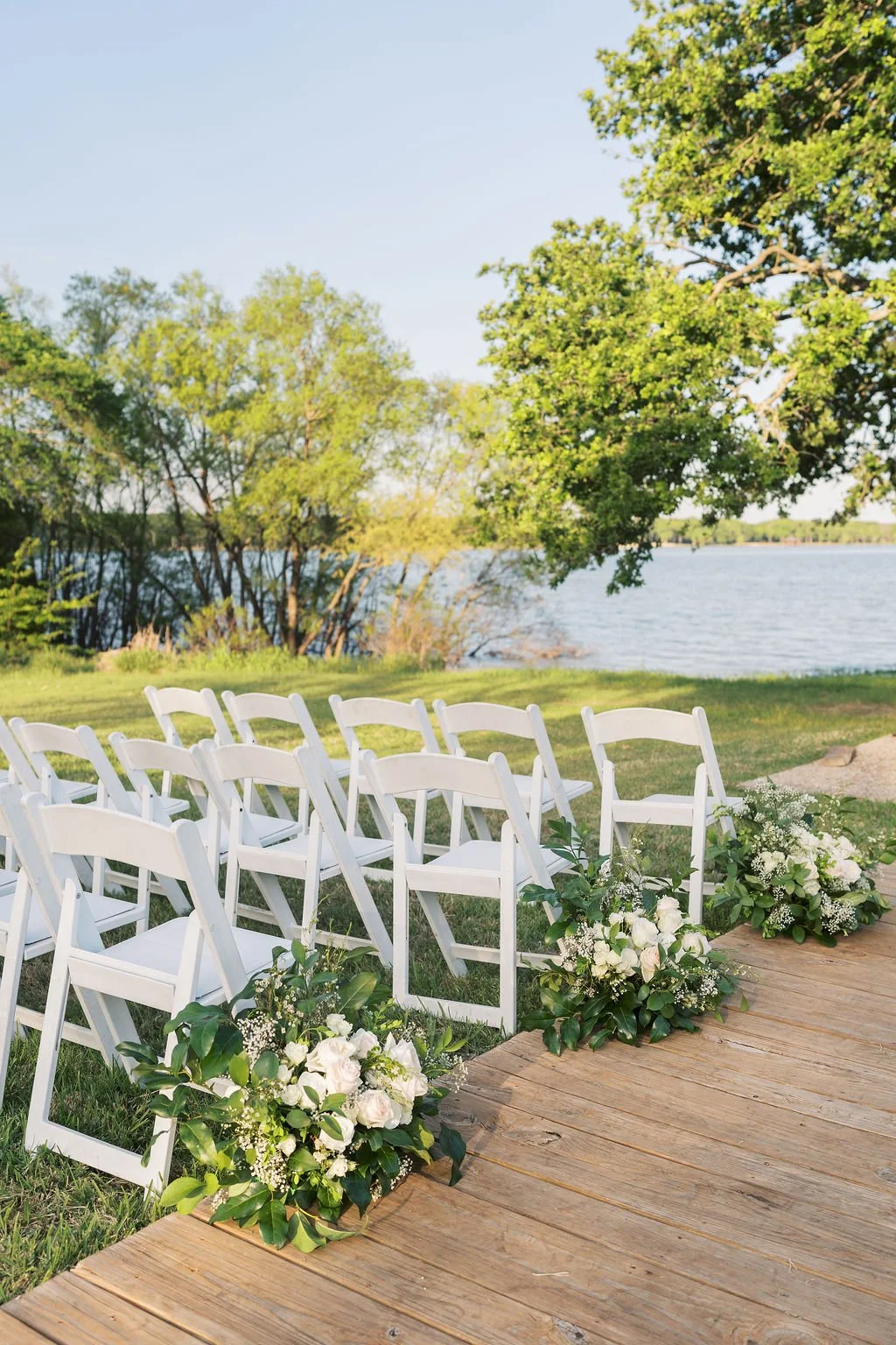 Outdoor wedding ceremony at Peninsula Ranch in DFW with white chairs arranged on a grassy area by a lake, decorated with white flowers and greenery, with trees and water in the background.