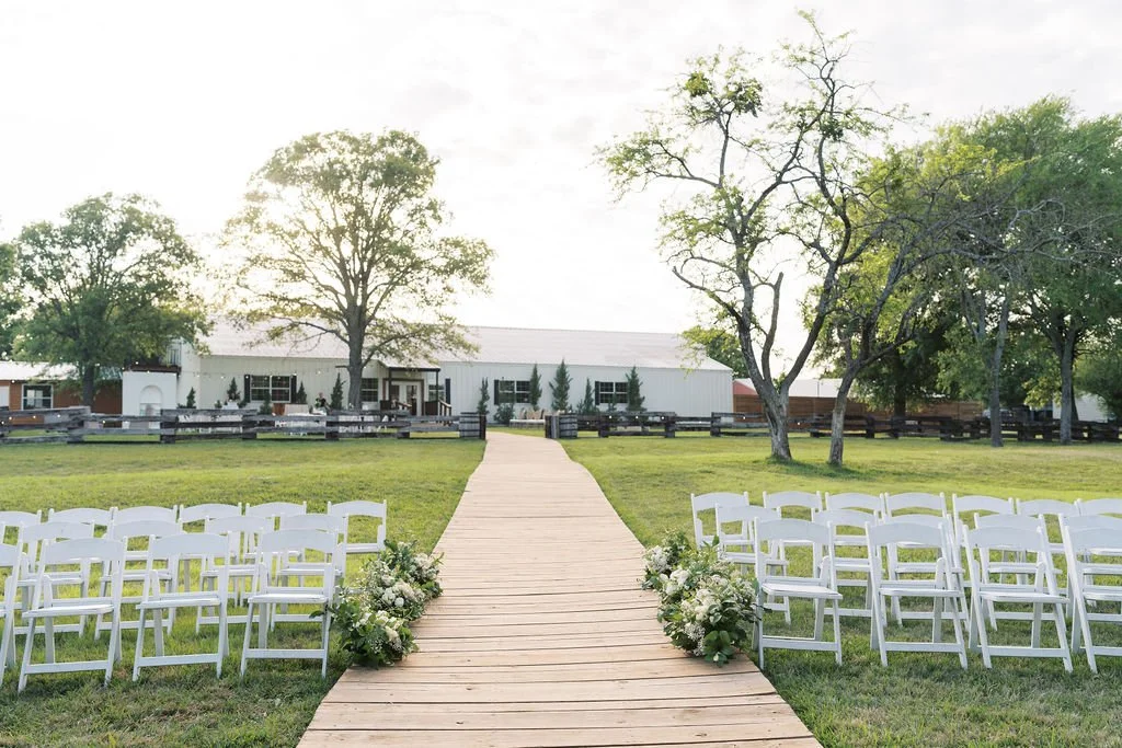Outdoor wedding ceremony setup at Peninsula Ranch in DFW with white chairs along a wooden aisle decorated with IZALENA floral arrangements, on a grassy field with trees and a building in the background.