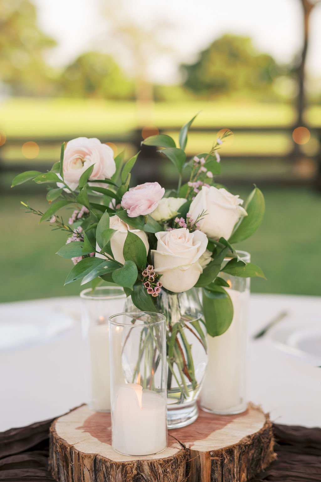 An IZALENA floral arrangement with white roses and pink flowers in a glass vase, placed on a wooden slice with candles around it, set outdoors at Peninsula Ranch Wedding Venue in DFW. 