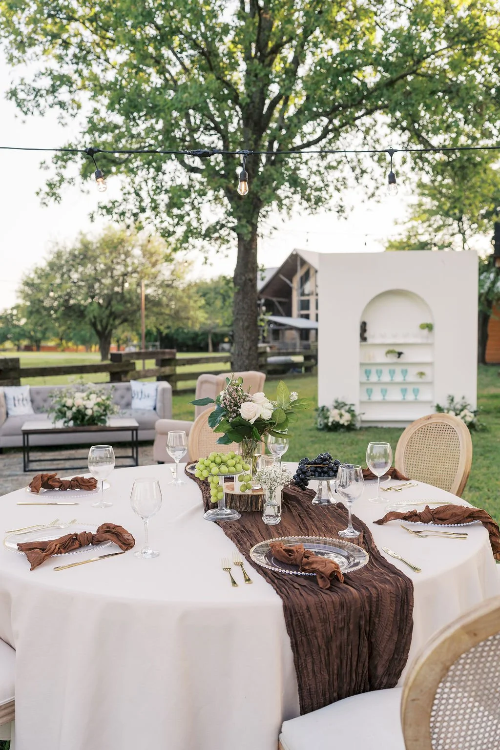 Outdoor table at Peninsula Ranch in DFW.with a white tablecloth, brown napkins, glassware, and IZALENA floral centerpiece, surrounded by wicker chairs, with a background of trees and a white shelf with glasses and plants.