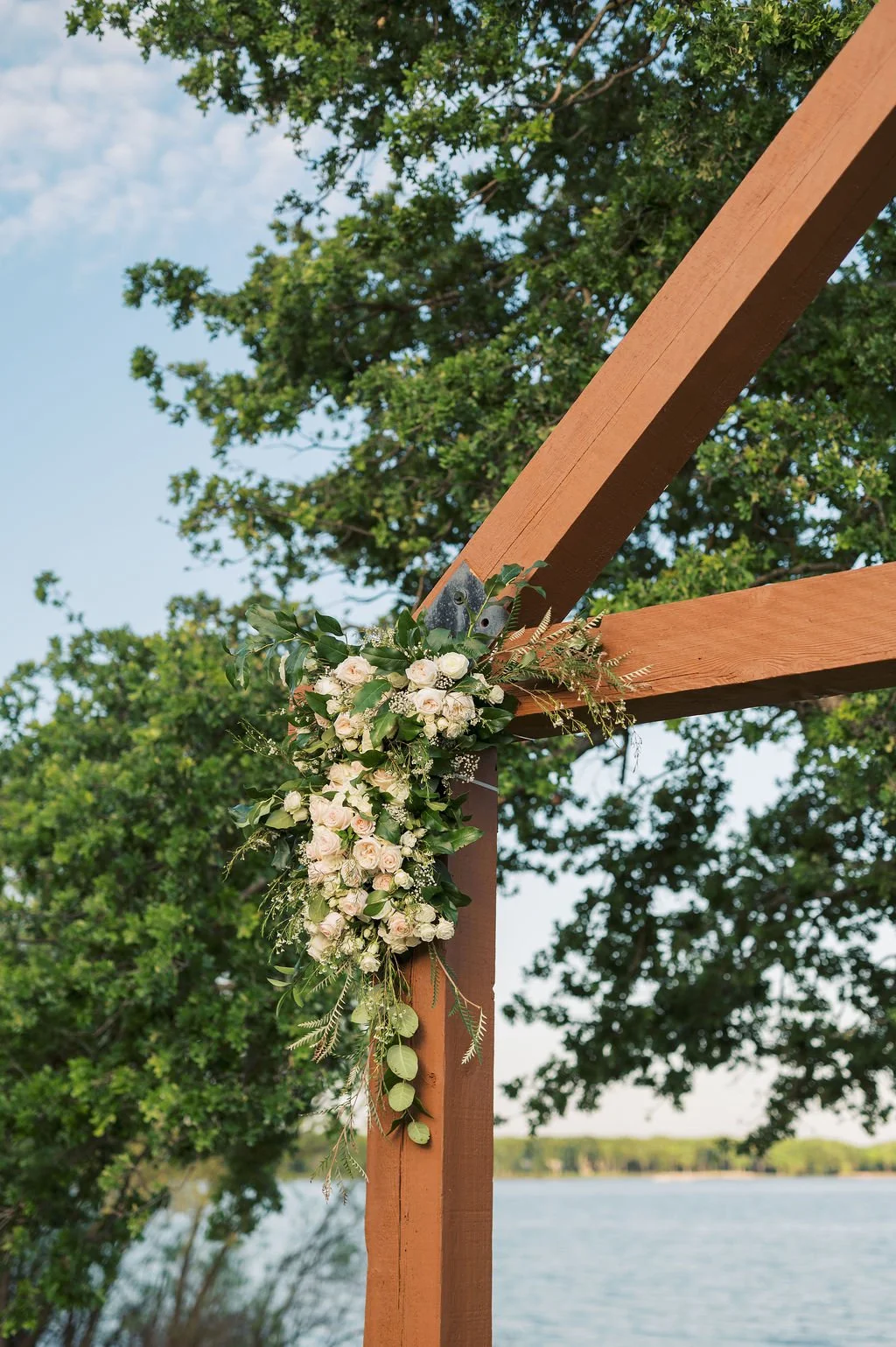 Decorative wooden arch with a IZALENA Floral House floral arrangement of white roses, green leaves, and other greenery, set outdoors near Lake Tawakoi at Peninsula Ranch in DFW.