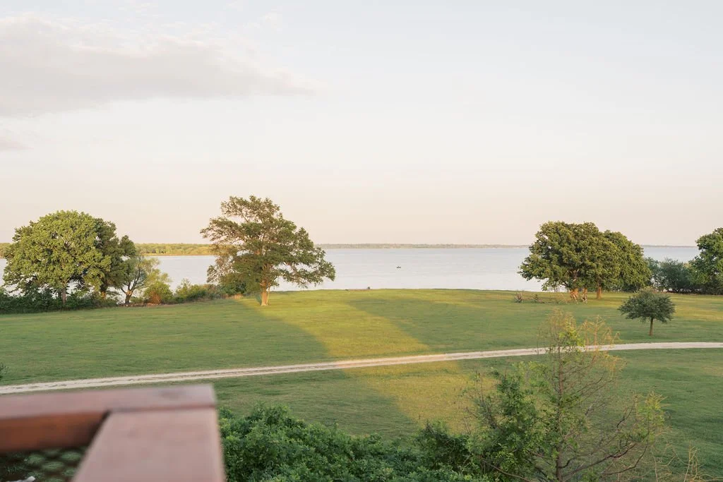 A scenic view of a grassy area with trees near a body of water, under a partly cloudy sky, with a wooden railing in the foreground at Peninsula Ranch in DFW 