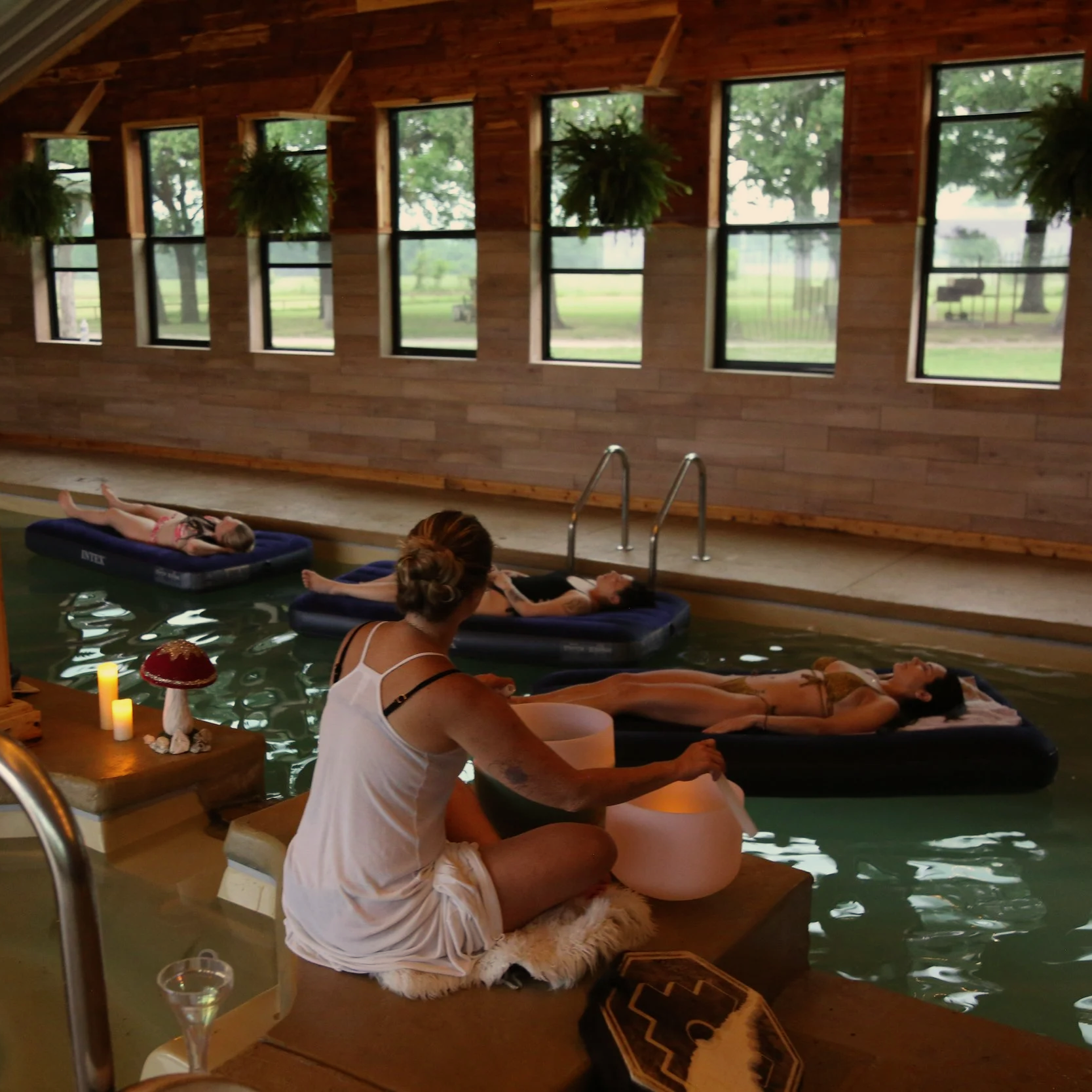 Indoor pool with wooden walls and large windows, showing outdoor trees and greenery; three women are floating on inflatable loungers in the pool, and a woman dressed in white is sitting on a platform by the pool, playing a crystal singing bowl.
