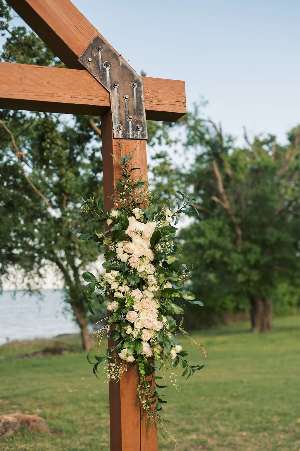 A wooden wedding arch decorated with white roses, greenery, and small white flowers outdoors on Lake Tawakoni at Peninsula Ranch in DFW.