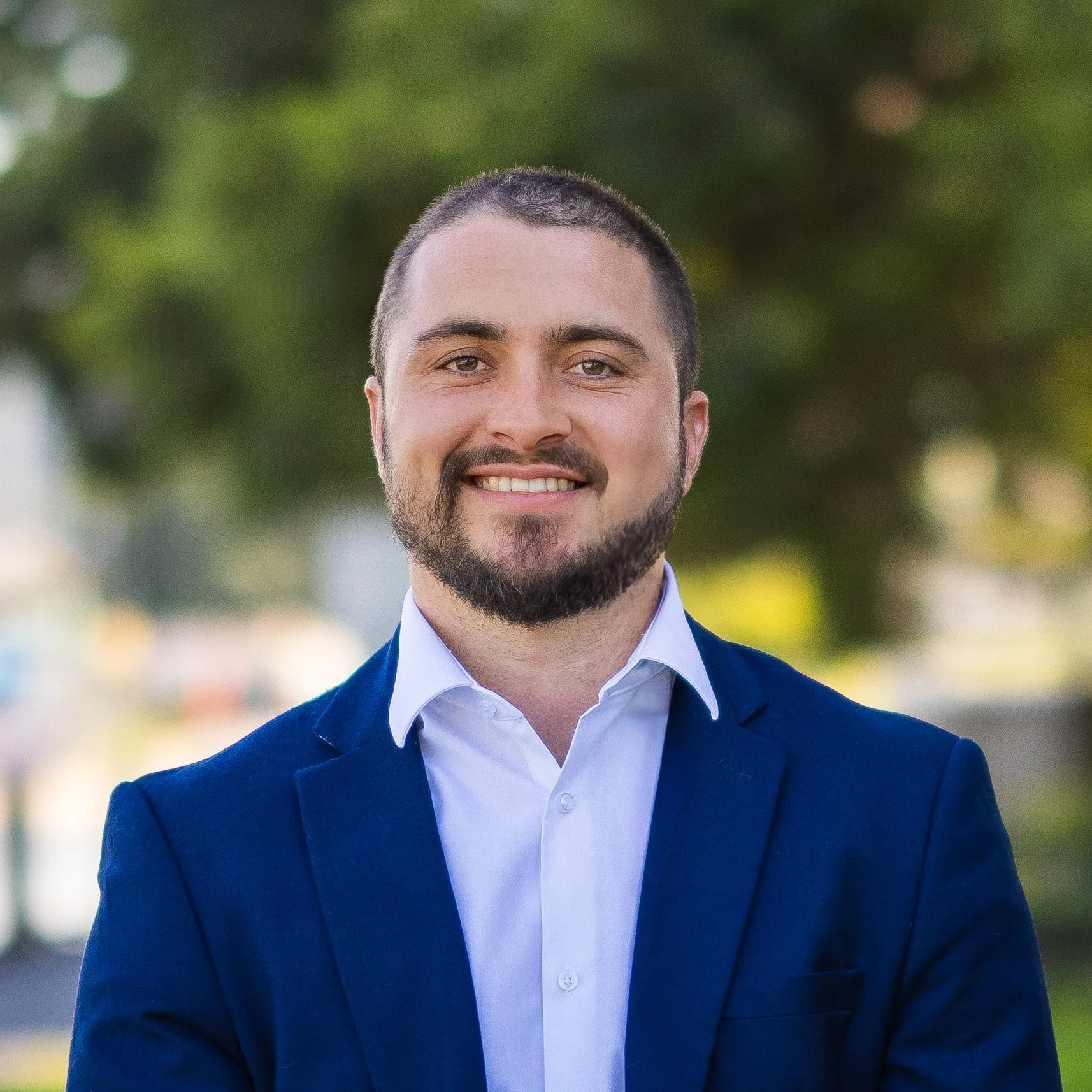 A young man with short dark hair and a beard, wearing a white shirt and a navy blue blazer, smiling outdoors with trees in the background.