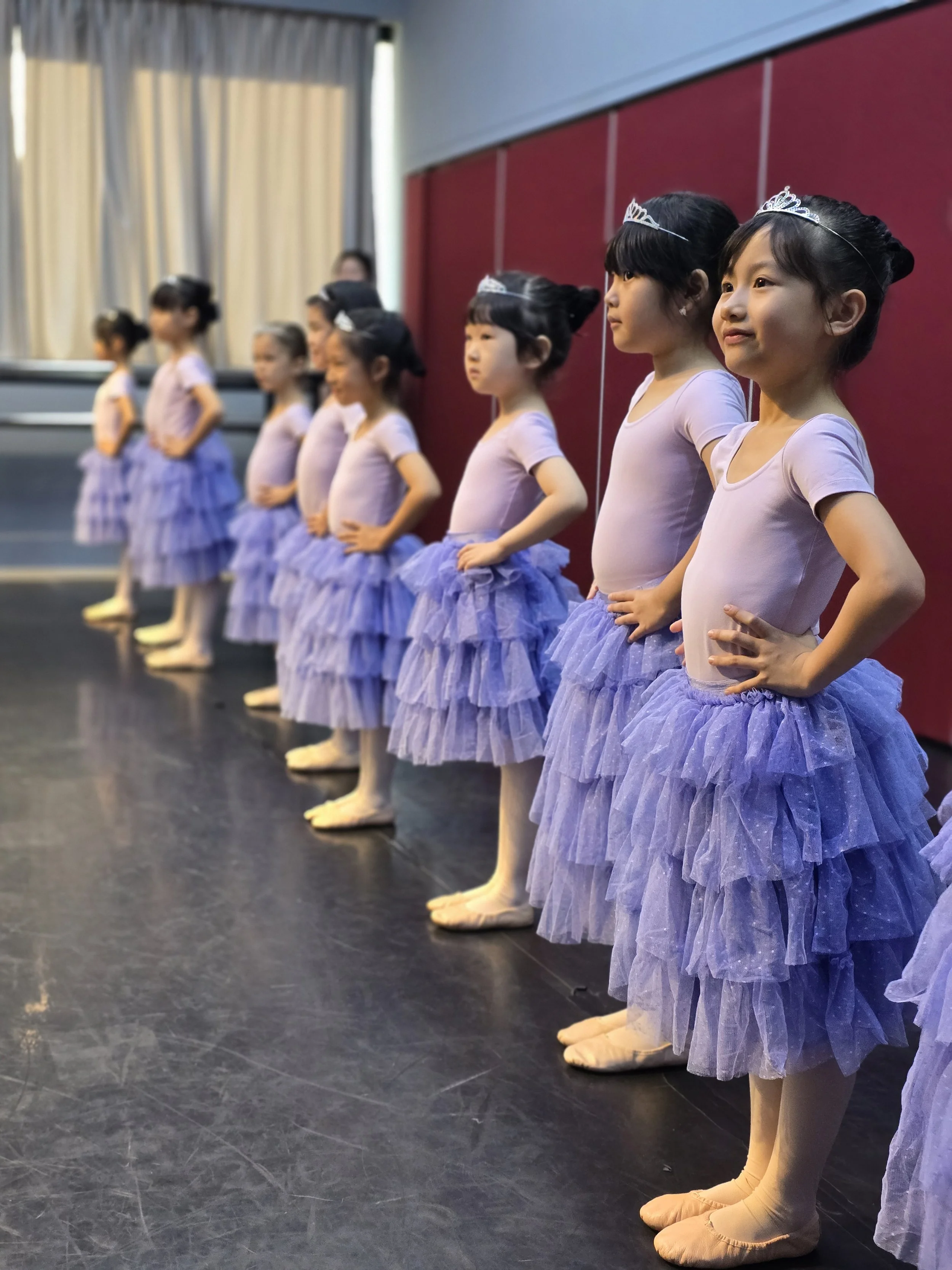Young girls dressed as ballet dancers in lavender tutus and crowns, standing in a line on stage during a dance recital.