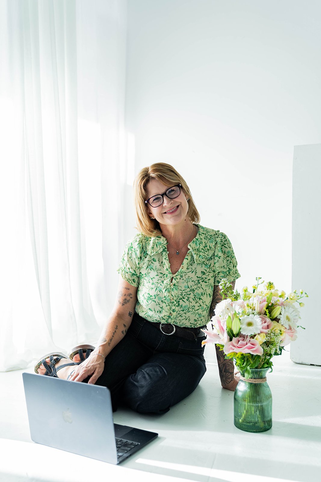 A woman sitting on the floor by a laptop, smiling, with a vase of flowers nearby in a bright, minimalist room.