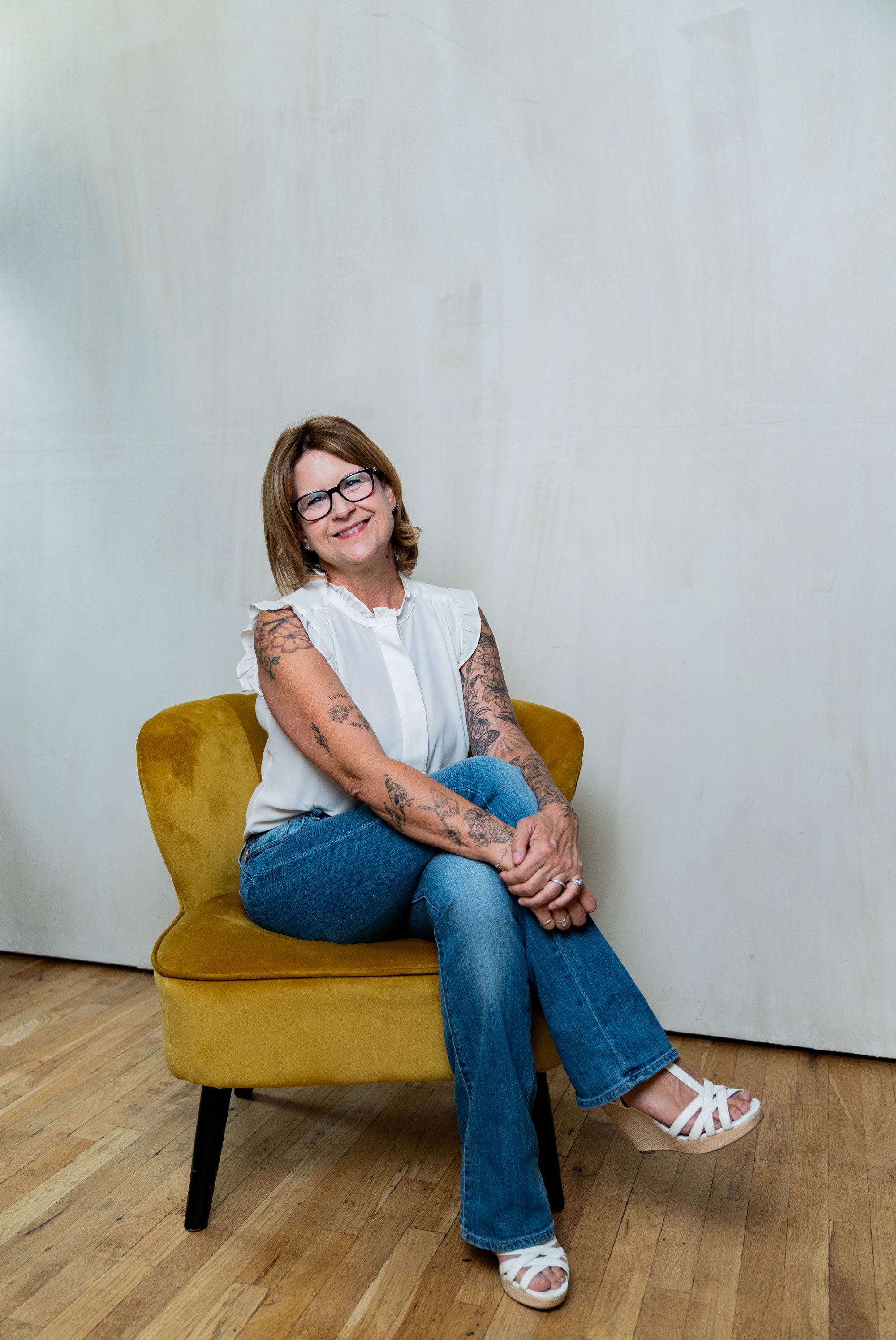 A smiling woman with glasses and tattoos, sitting on a yellow upholstered chair with black legs in a room with wooden floors and a plain off-white wall, wearing a white sleeveless blouse, blue jeans, and white strappy wedge sandals.