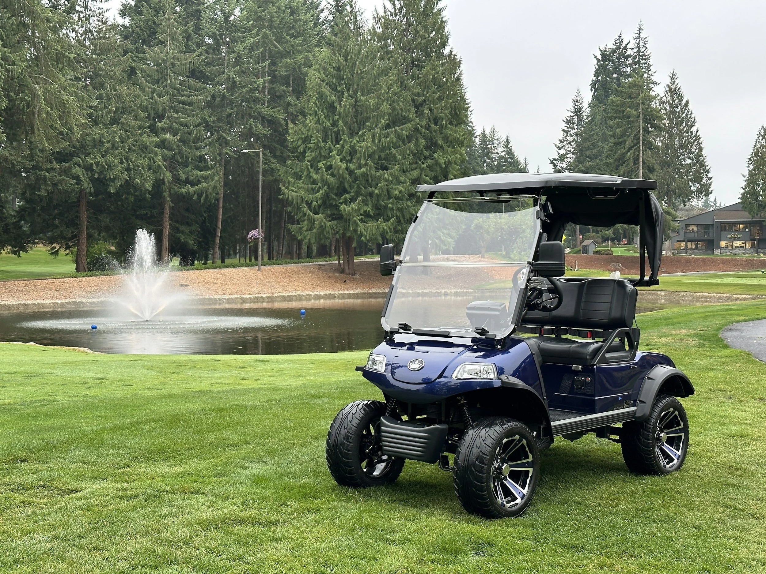 A blue golf cart parked on a well-manicured golf course near a pond with a fountain, surrounded by tall trees and some residential buildings in the background.