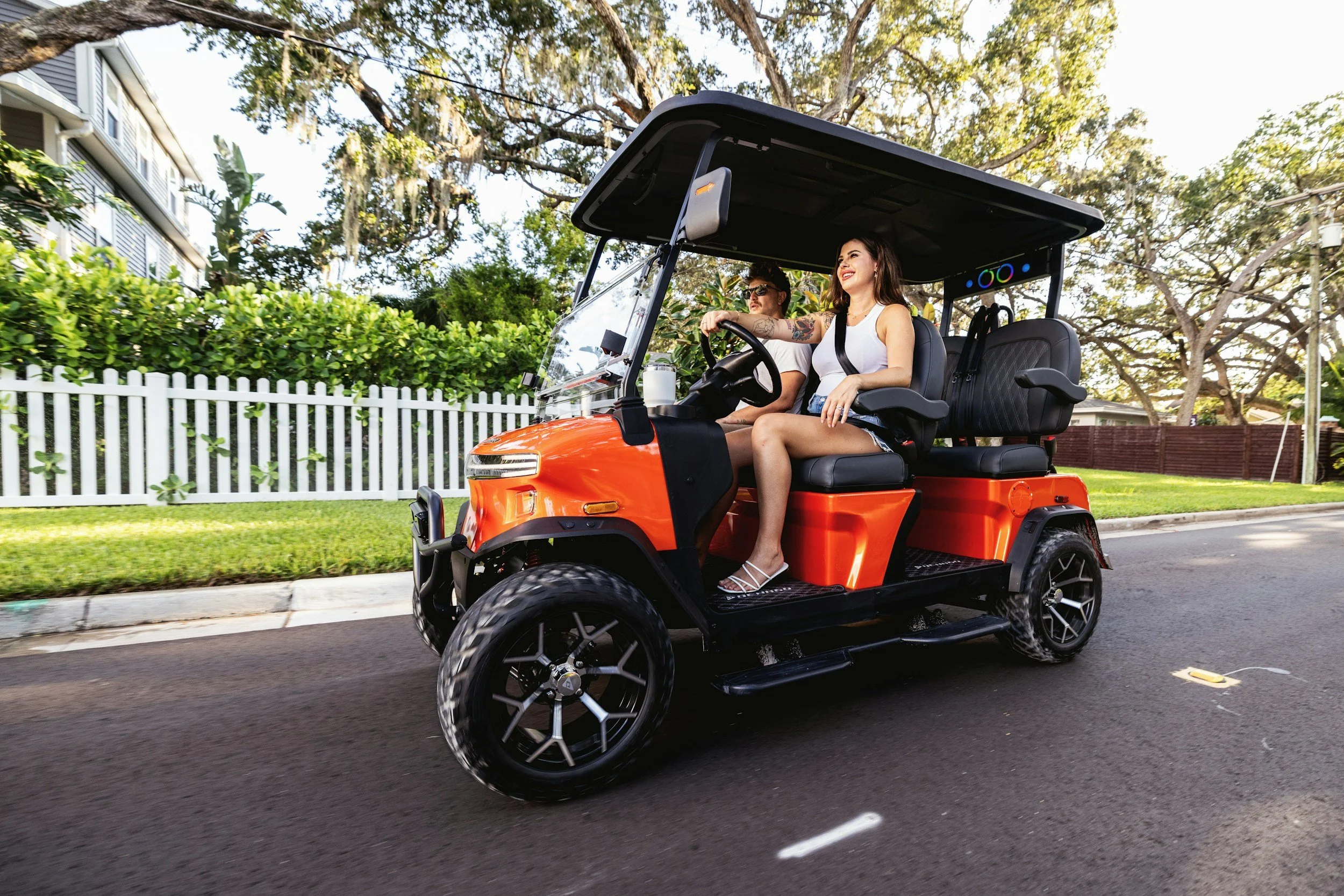 Two women riding in an orange golf cart on a suburban street with green trees and a white picket fence in the background.