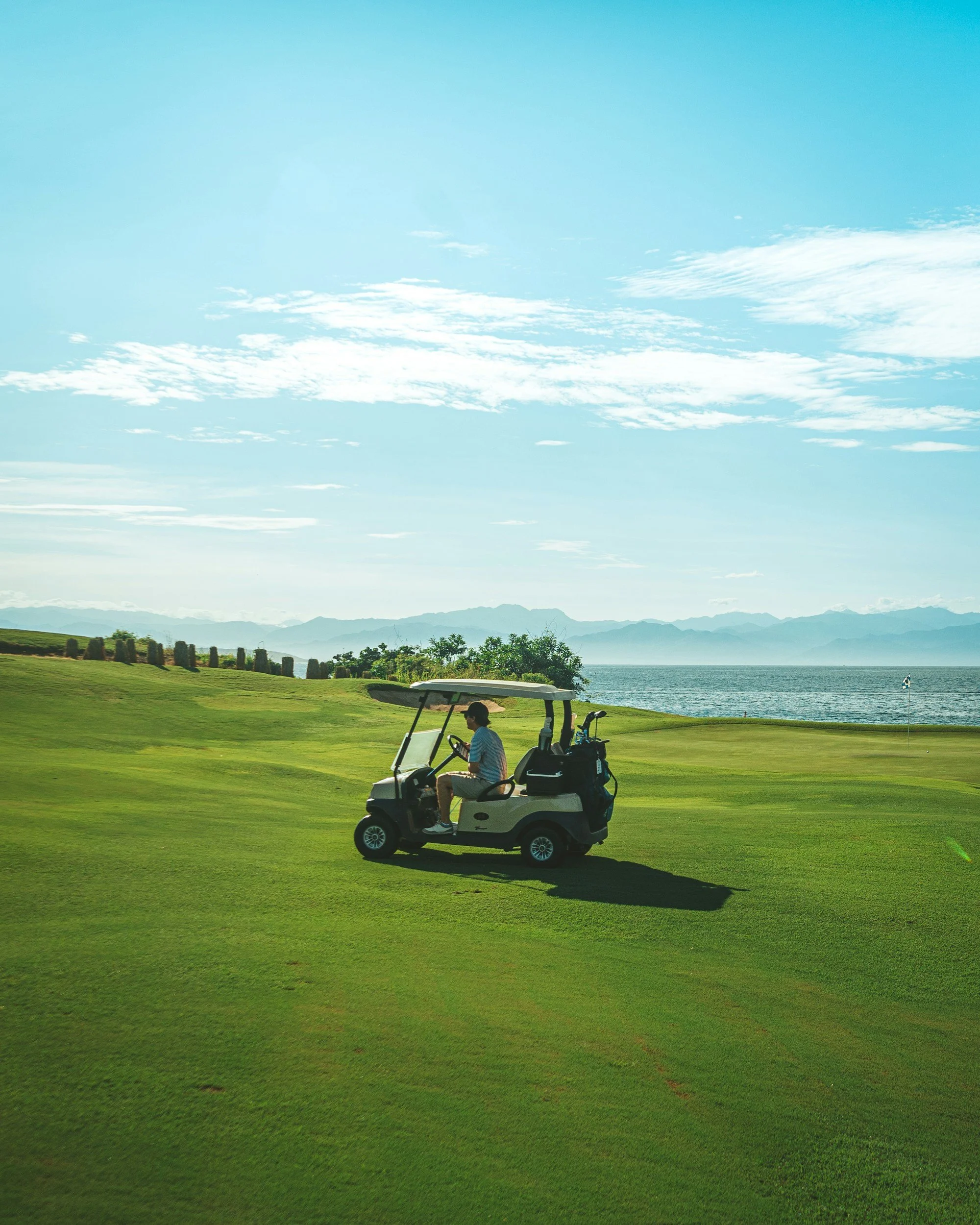 A person riding a golf cart on a lush green golf course near a body of water with mountains in the background under a partly cloudy sky.