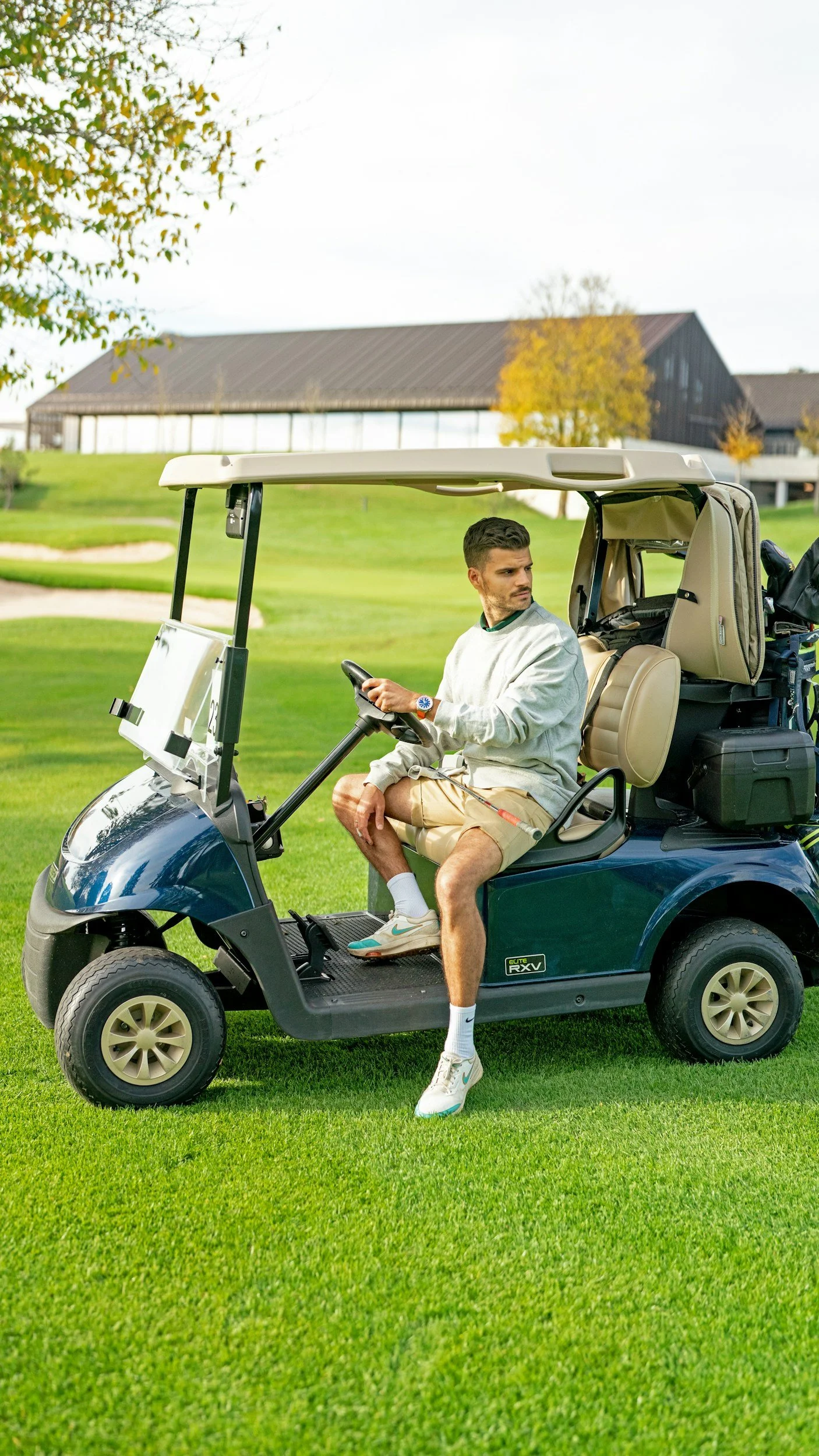 A young man sitting in a golf cart on a golf course, holding the steering wheel and looking to his left. The golf cart is parked on green grass with trees and a barn-style building in the background.