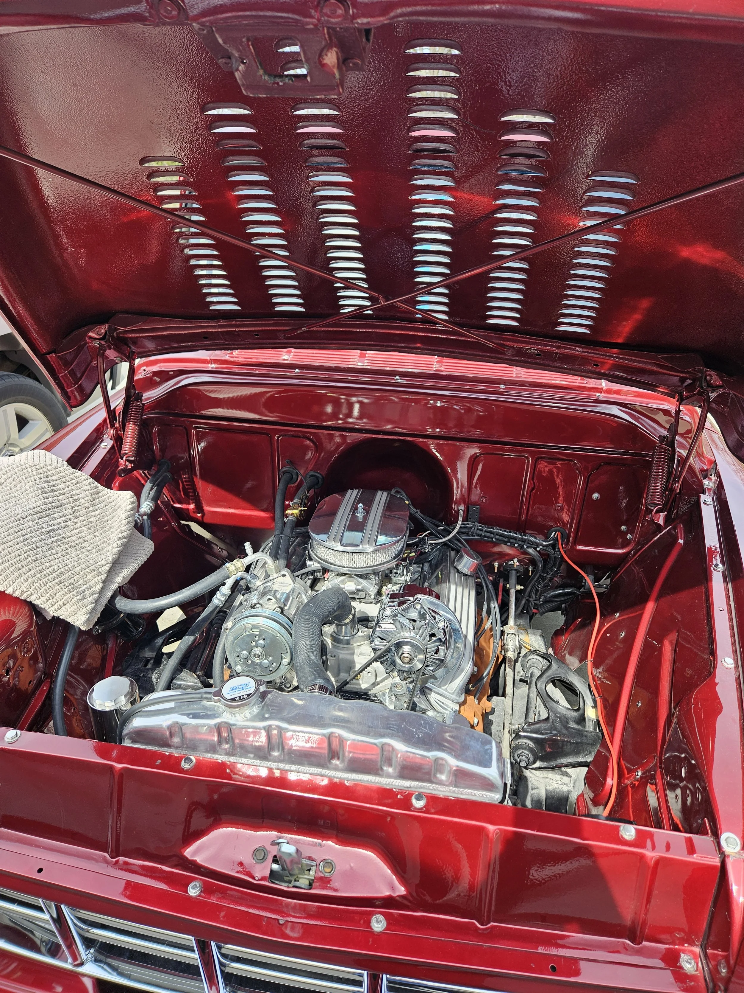 View of a classic red car engine bay with the hood open, showing a polished supercharger, engine components, and a red-painted interior, including the hood underside with ventilation slots.