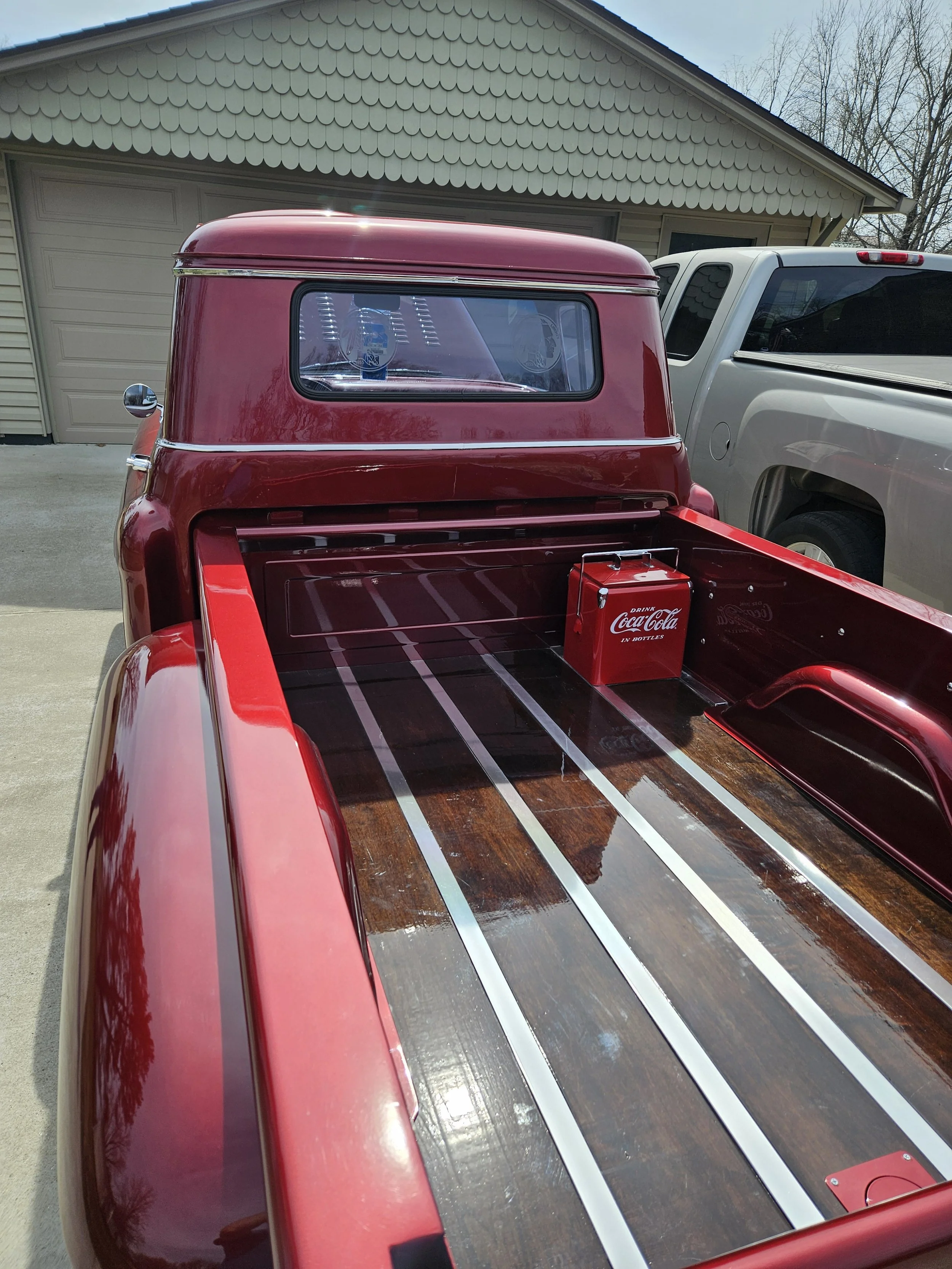 A red vintage pickup truck with a wooden bed and a Coca-Cola box inside, parked in front of a house with a beige garage door and siding, and a white pickup truck parked beside it. There are leafless trees in the background.